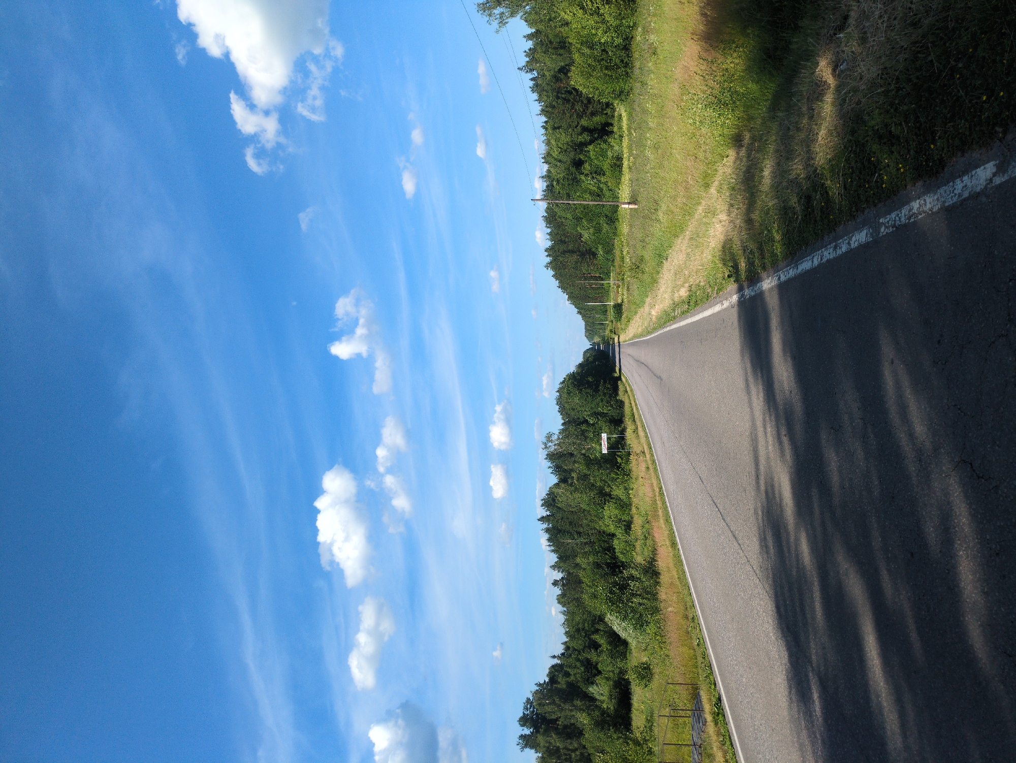 This is a landscape photograph taken from a low vantage point, possibly from a moving vehicle, showing a rural road in Velet'ma, Russia. The image is oriented vertically, with the sky dominating the left half of the frame and the road and its surroundings on the right. The sky is a vibrant blue with scattered white cumulus clouds, indicating fair weather. The time of day appears to be daytime, likely late morning or early afternoon, given the bright sunlight and distinct shadows cast on the road.
The road itself is asphalt and appears to be a two-lane country road, with a white line marking the edge on the left side. The right side of the road is bordered by a grassy verge and a dense line of green trees. The trees form a thick forest, with the foliage appearing lush and healthy. Further down the road, the forest seems to extend as far as the eye can see. There are utility poles with wires running along the side of the road, suggesting that this is a settled area. A small white sign is visible in the distance, mounted on a pole at the edge of the forest, but the text on it is illegible. The overall impression is one of a peaceful, rural setting on a clear, sunny day. The scene evokes a sense of journey or travel through the countryside.