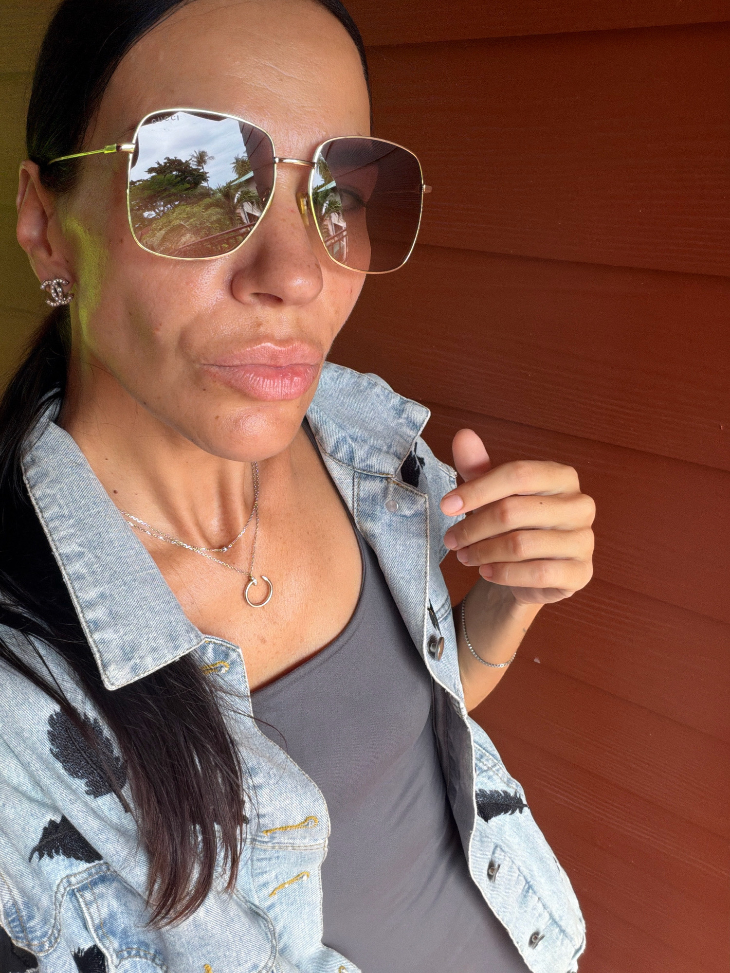A woman wearing large, gold-rimmed sunglasses, a denim jacket with black feather embroidery, and a grey tank top poses against a reddish-brown wooden wall. She is wearing a delicate silver necklace and a smaller silver chain with a circular pendant, and a diamond-encrusted Chanel earring. Her hand is raised, giving a thumbs up gesture. The background suggests an outdoor tropical environment, possibly in Ko Samui District, Thailand, with lush greenery reflected in her sunglasses.
