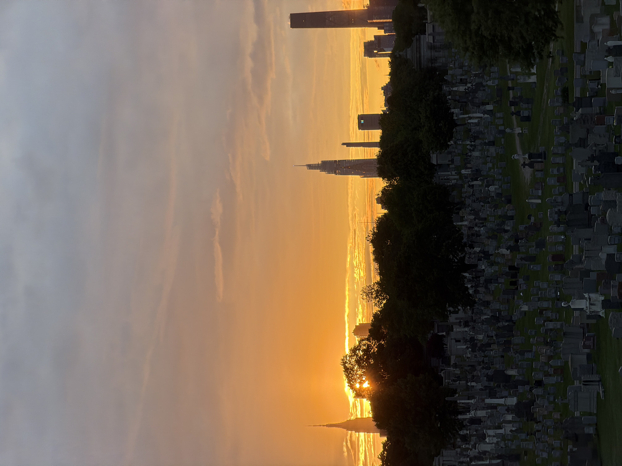 The image shows a golden sunset over a city skyline, viewed from a graveyard. The graveyard is located on a hill, with rows of tombstones visible. The skyline includes several skyscrapers, including the Chrysler Building and One Vanderbilt. The sunset is casting a warm glow over the city and the graveyard. The location is Green-Wood Cemetery in Brooklyn, New York.