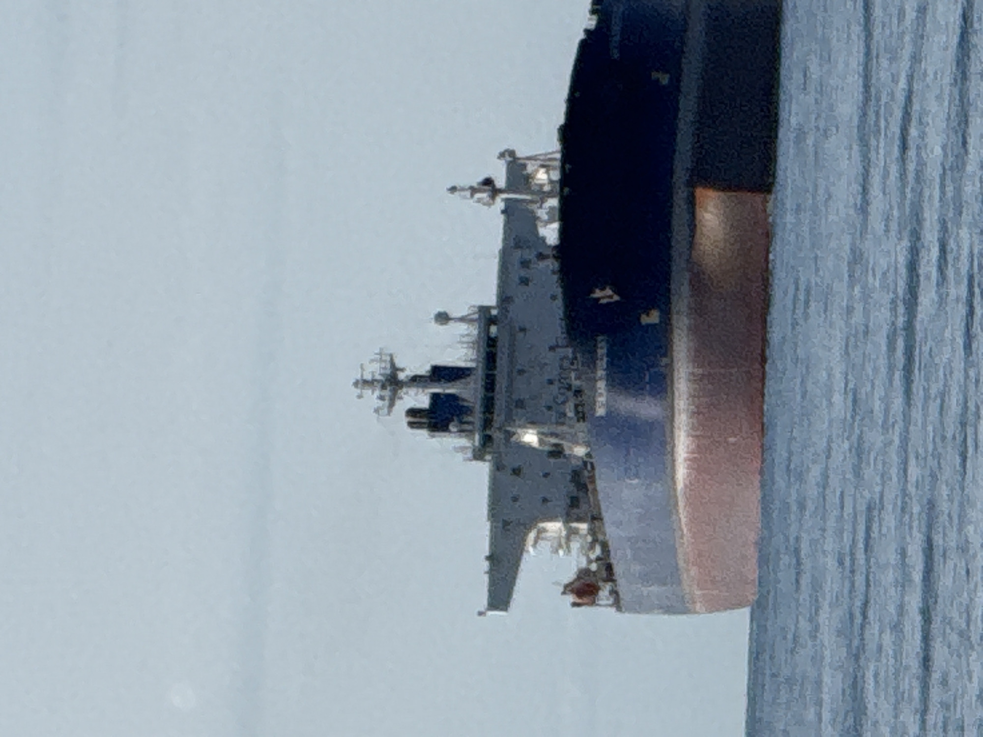 A large, dark blue cargo ship with a red hull is sailing on a calm, blue ocean under a pale blue sky. The ship is the primary subject, filling most of the frame. It features a prominent superstructure with several levels, antennas, and radar equipment. The ship's name, partially obscured by glare, appears to be "CORAL". The bow of the ship is visible on the right side of the image. The water is calm with subtle ripples. The image is a photograph taken from a distance, likely from another vessel or the shore. It is difficult to determine the exact location within Teriberka, Russia, without further context.