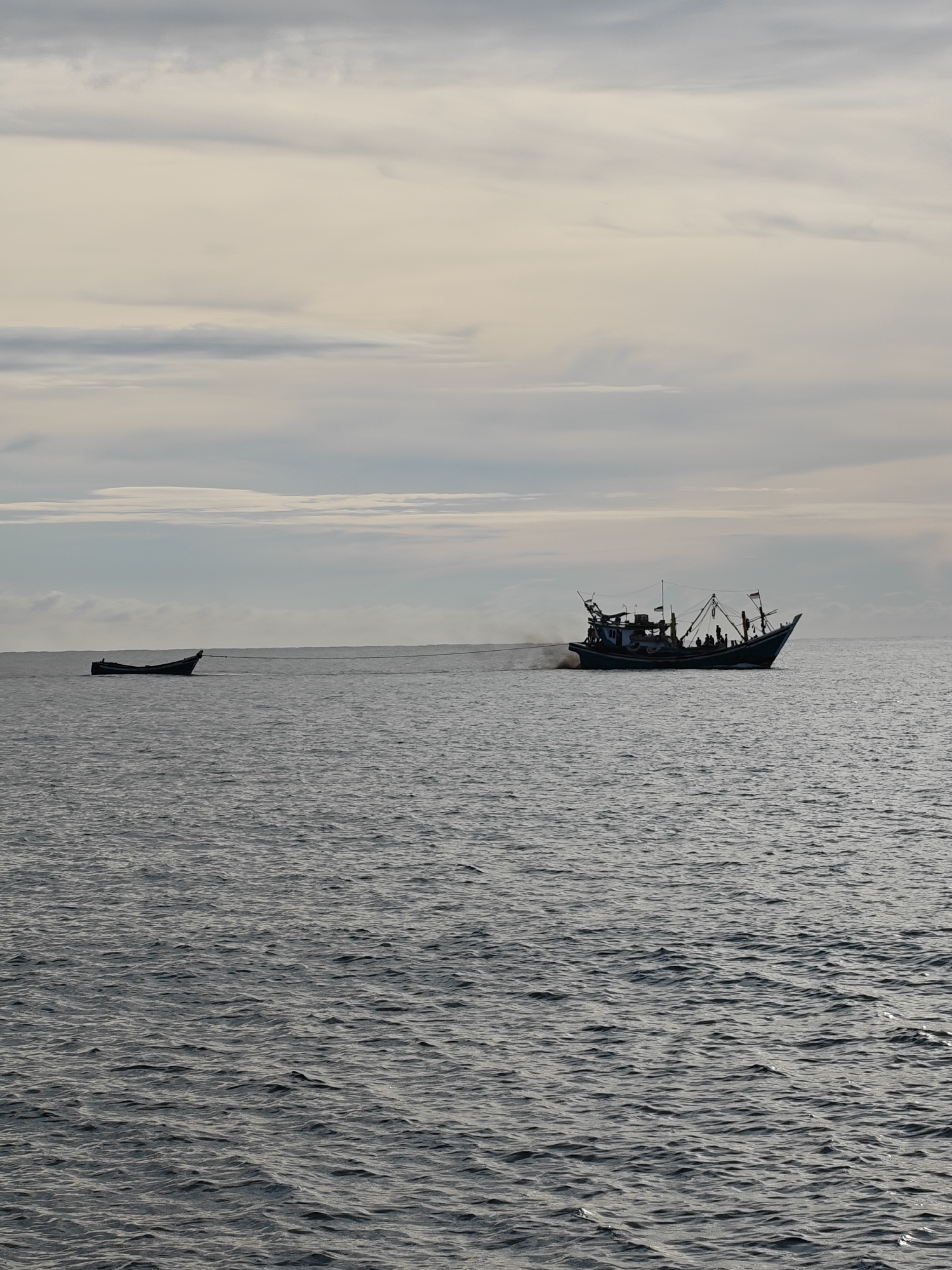 The image depicts a coastal scene, likely in Lhokseumawe, Indonesia, featuring two boats on the water under a sky with muted clouds. The larger boat, positioned on the right side of the frame, appears to be a fishing vessel with a visible crew and associated equipment like nets and masts. The boat emits smoke. To the left, a smaller boat is towed by a rope, and its occupants, if any, are not clearly visible. The water shows moderate waves. The scene is captured in a way that renders the details of the boats and sky silhouetted.