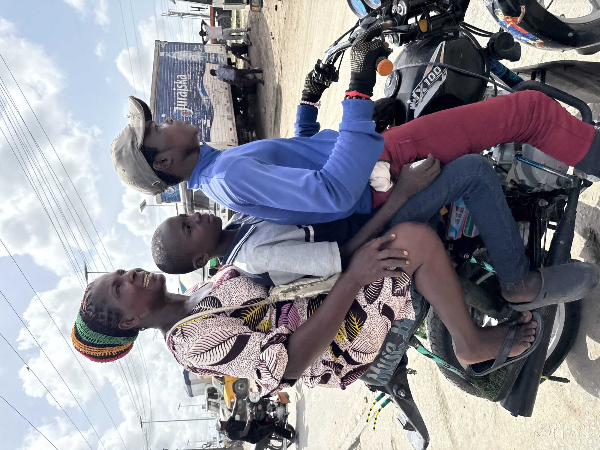 A woman, a boy, and a man are riding on a motorcycle. The woman is seated in the back, wearing a patterned dress, a crossbody bag, and a colorful knitted hat. The boy is in the middle, wearing a grey sweater. The man is driving, wearing a blue sweatshirt, red pants, a cap, and gloves. The motorcycle is a black "JX100" model with a sticker of a soccer player on the gas tank. The environment appears to be an urban street, with a truck and other motorcycles visible in the background. The truck has “Jurajska” written on its side. The location is likely Lekki, Nigeria.
