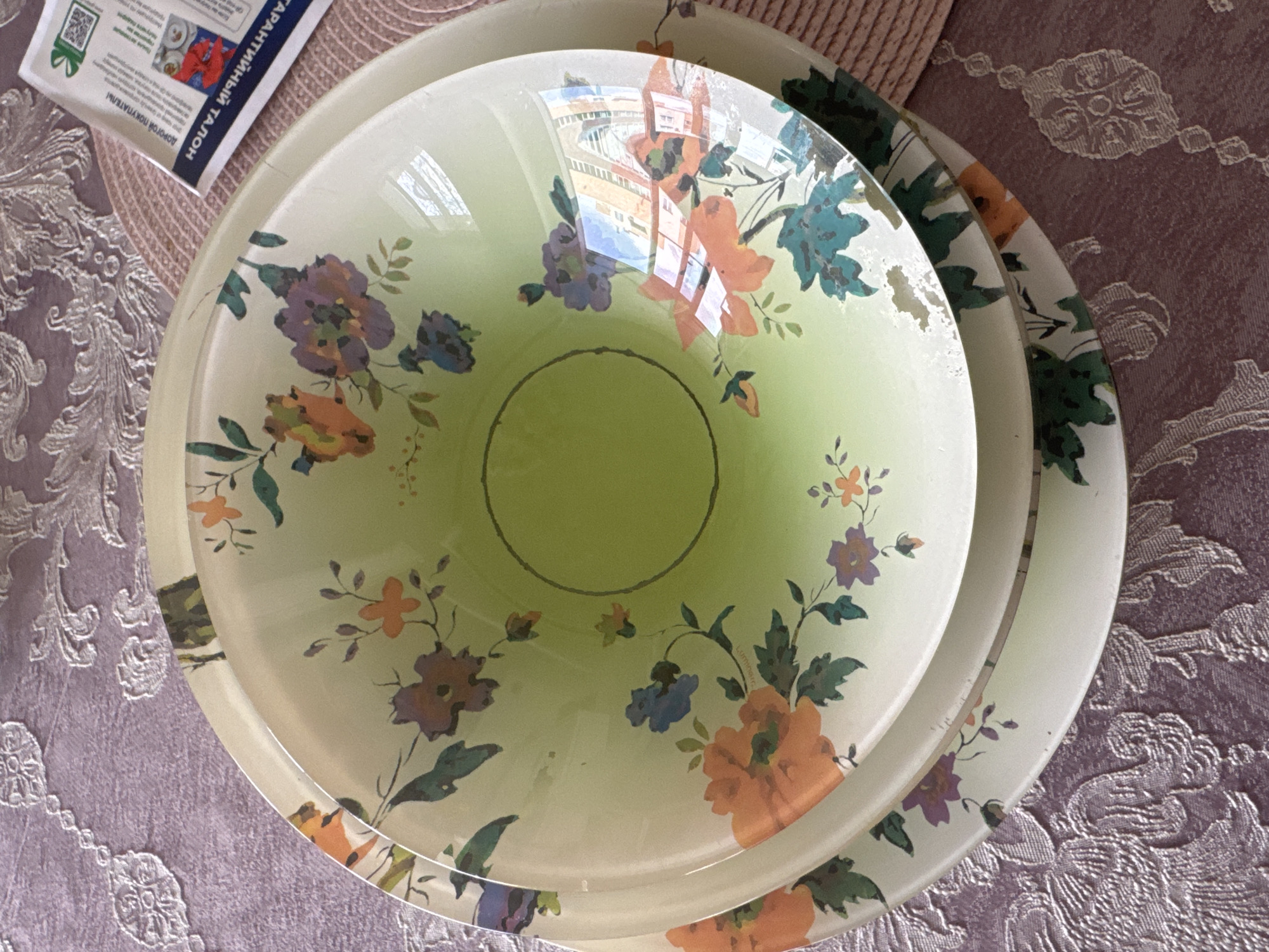 A stack of three decorative bowls with floral patterns is placed on a textured purple tablecloth. The top bowl is larger and appears to be a serving bowl with a lime green interior and an orange and purple floral design on the exterior. The other two bowls are smaller and stacked beneath it, with similar floral patterns. A paper document with text in Russian and a QR code is partially visible on the right side of the image, placed on a pink woven placemat. The background is a detailed view of the floral patterned tablecloth and a hint of a white decorative trim. No people are visible in the image. The location is described as Unknown, Russia.
