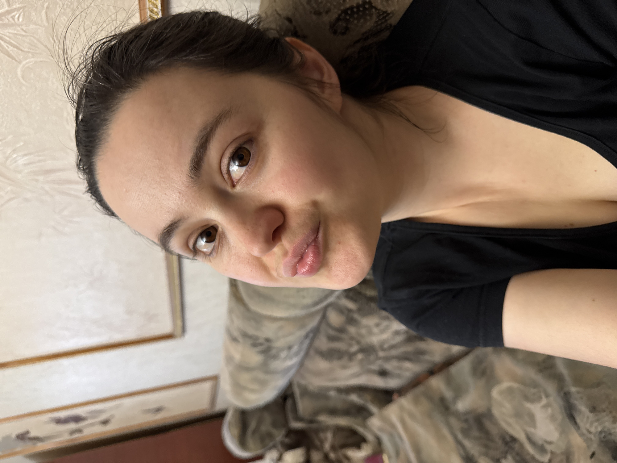 A close-up, rotated portrait features a young woman indoors. She has dark hair, pulled back with some strands loose around her forehead, and warm brown eyes that meet the camera's gaze. Her lips are slightly pursed in a playful or affectionate expression. Her complexion is natural, with a hint of rosiness on her cheeks. She is dressed in a plain black t-shirt, visible around her neck and shoulders.
The background suggests an indoor setting, likely a room in a home. A light-colored wall with a subtle decorative pattern, possibly wallpaper, and a wooden frame or molding with golden accents are visible. Part of a patterned piece of furniture, which appears to be a sofa or bed, is also in the frame. The lighting is soft and even, indicating either natural daylight from an unseen window or ambient artificial lighting. The overall mood is casual and intimate. There is no visible text in the image. The location context provided is Izhevsk, Russia.