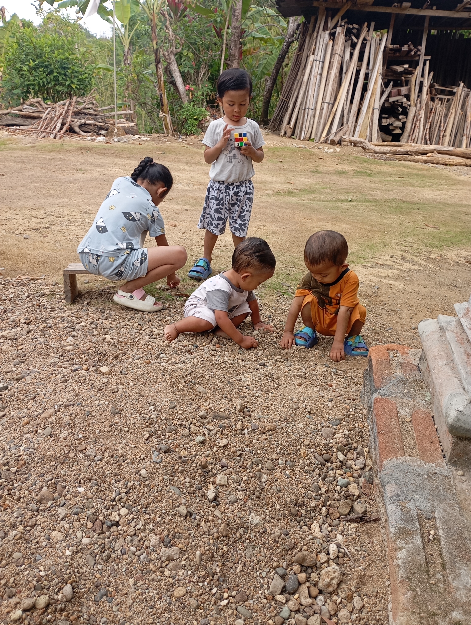 In Ketepung, Indonesia, four children are playing on a gravelly ground in an outdoor setting. One child, a boy wearing a grey t-shirt and patterned shorts, stands holding a Rubik's Cube. To his left, a girl in a blue pajama set with panda motifs is crouched on a wooden stool, her hair in a braid. In front of her, two younger children are also on the ground. One, a boy in a grey and white striped shirt and grey shorts, is looking down at the ground. Beside him, a boy in an orange shirt and orange shorts, wearing blue sandals, is also looking down. Behind the children, a pile of logs and a rudimentary wooden structure are visible, suggesting a rural or undeveloped area. The ground is covered in small pebbles and dirt, with some vegetation and trees in the background.