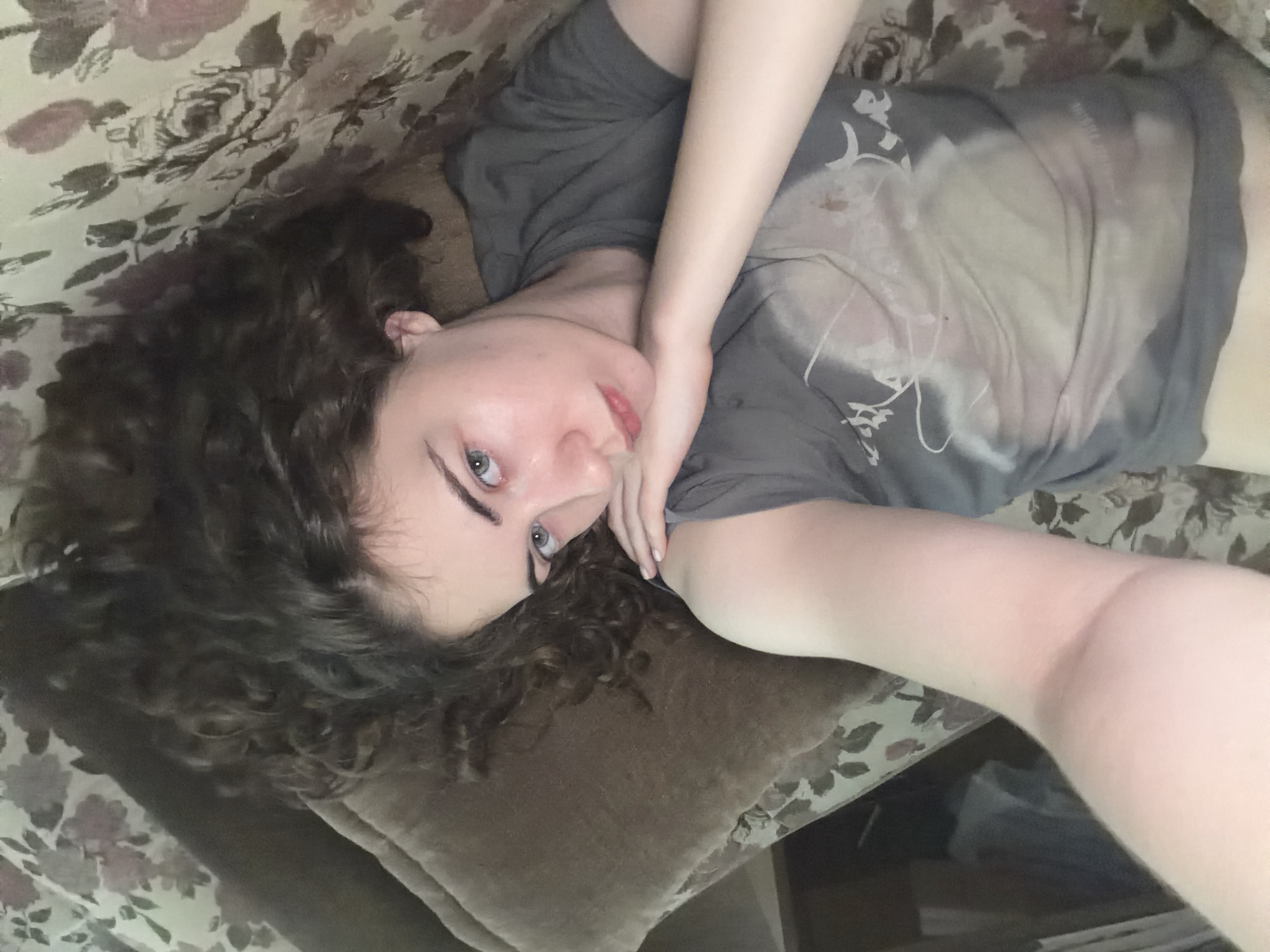 A young woman with curly brown hair and blue eyes lies on her side on a floral-patterned couch, looking towards the camera. Her right arm is bent, and her hand rests on her cheek, supporting her head. She is wearing a dark gray t-shirt. The background consists of a brown pillow and the couch with a floral print. The lighting appears to be natural. The image was taken in Zavodoukovsk, Russia.