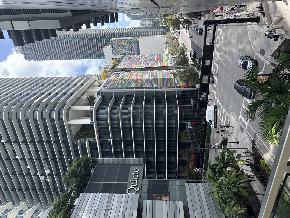 This image captures an elevated daytime view of a bustling urban intersection in the Brickell neighborhood of Miami, United States of America.
In the foreground, a gray metal railing and blurred green palm fronds suggest the vantage point is from a balcony or upper floor of a building.
The left side of the frame is dominated by a tall, modern skyscraper featuring numerous floors with curved white balcony railings and dark glass windows. Green foliage is visible on several lower terraces. Below this, a multi-story building segment displays a prominent "Quinto" sign in white lettering on a dark gray facade. A digital display screen within this segment shows a partially visible image of a person.
Towards the middle and right, a distinct multi-story building features a vibrant, multi-colored striped facade composed of horizontal and vertical panels in red, orange, yellow, green, blue, and purple hues. Above this building, a large, colorful circular abstract mural is affixed to a white wall. Further into the background, multiple tall, slender skyscrapers, primarily gray with repetitive window patterns, rise against a sky with scattered white clouds.
At street level, an asphalt intersection with white crosswalks and lane markings is visible. Overhead, black traffic light gantries span the intersection. A white cargo van is positioned on a crosswalk, facing right, with a black SUV stopped behind it. Another silver SUV is further back, appearing to turn right. On the street extending into the distance, several other vehicles are present. On the left sidewalk, two bicycles are parked near a rack amidst palm trees and greenery.
Pedestrian activity is moderate: a group of approximately seven individuals is crossing a crosswalk on the left, moving from left to right. On the right sidewalk, a larger group of around fifteen pedestrians walks or stands. Three individuals are also visible walking towards the bottom right corner of the frame. The environment is well-lit by daylight, and numerous palm trees are interspersed along the sidewalks and medians.
