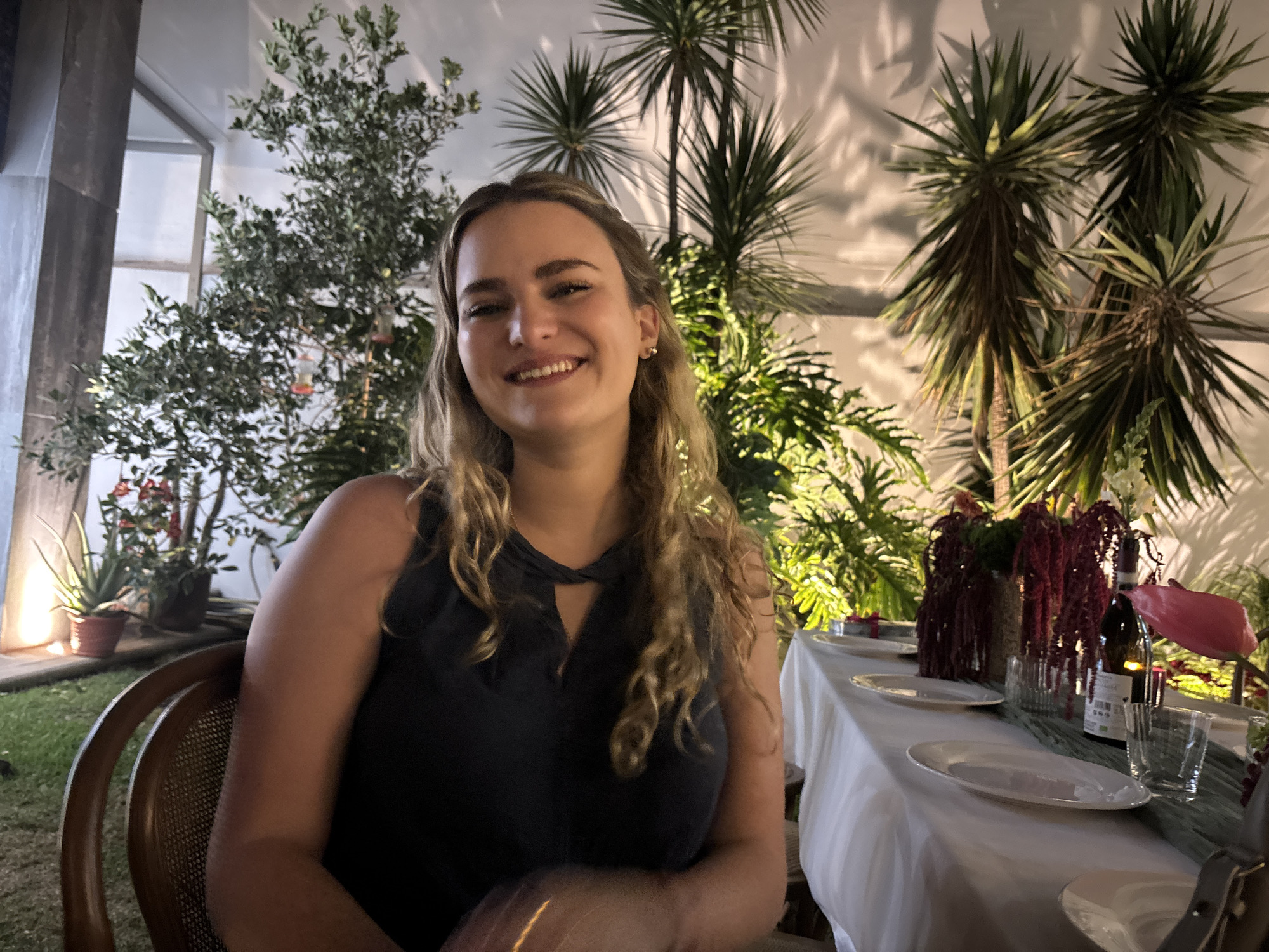 The image displays a nighttime outdoor or semi-outdoor social gathering area. A young adult female with light-colored wavy hair and a dark sleeveless top is centrally positioned, smiling directly at the camera. She wears a gold stud earring in her visible ear. To her right, a dining table is set with a white tablecloth, a dark green runner, multiple white oval plates, clear drinking glasses, a bottle of red wine, and a decorative centerpiece composed of deep red cascading plant material and a pink anthurium flower. The background features dense green foliage, including bushy trees with small leaves and small red hanging objects on the left, and several tall, spiky-leaved plants (possibly Yucca or Dracaena species) on the right, which cast pronounced shadows on a light-colored wall. Potted aloe vera plants and other flowering plants are visible near a ground-level light source on the far left. The woman is seated in a chair with a wicker-style back, partially visible on the left. The scene is illuminated by warm artificial lighting. The specific location within the city of Puebla, Mexico, is not discernible from the image content.