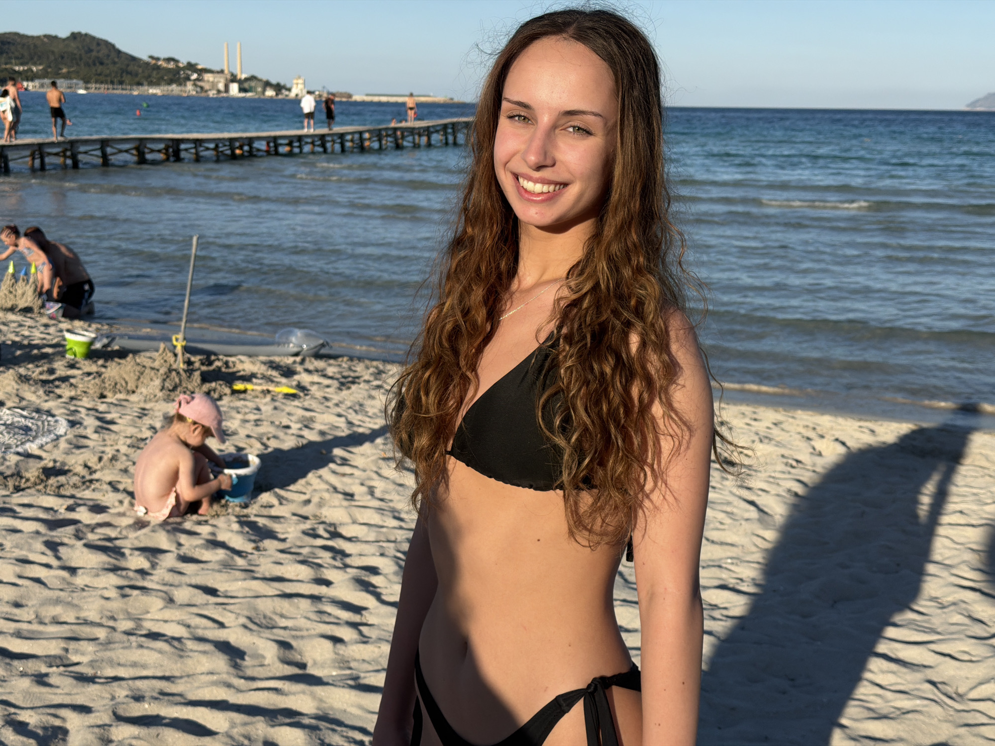The image is a medium shot of a woman smiling at the camera, wearing a black bikini, on a sandy beach. Her hair is long, wavy and brown. In the background, there's a group of people on a pier extending into the ocean. Further to the left, a child wearing a pink hat is playing in the sand with a blue bucket. In the distance, there is a hazy mountain range. This scene is in Alcúdia, Spain.