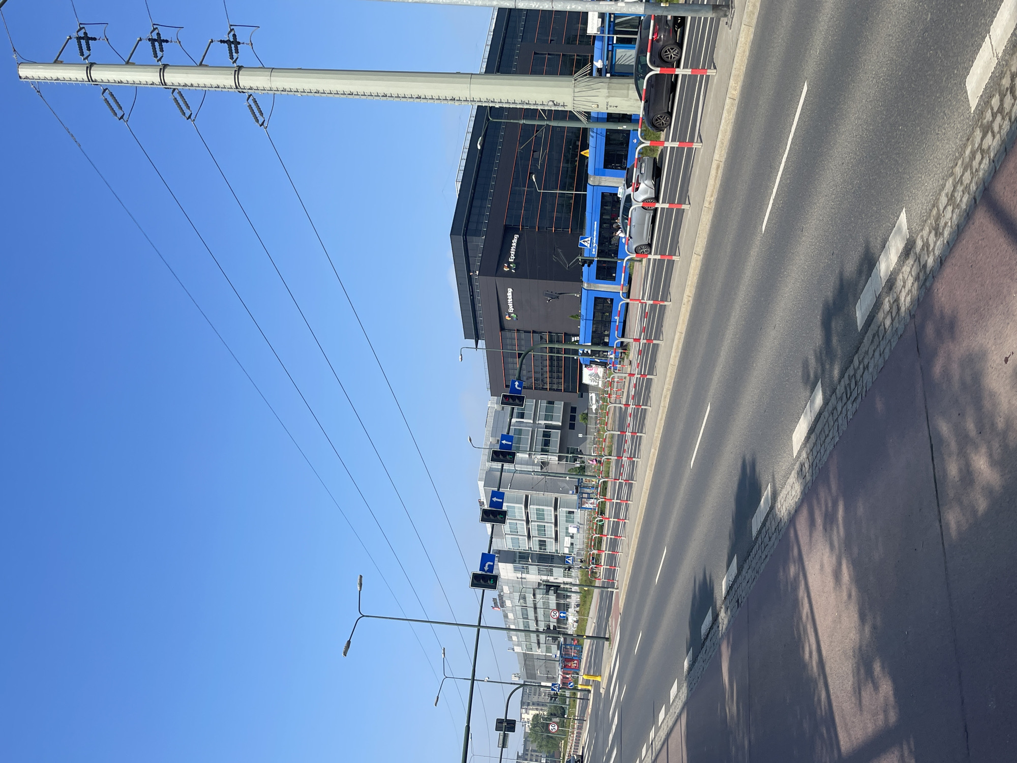 An urban street scene unfolds under a clear blue sky on a sunny day. The image, originally rotated, shows a multi-lane road running through the frame, with white lane markings and pedestrian crossings visible. To the right of the road, a sidewalk with grey pavers and a reddish-brown pedestrian or cycling path can be seen.
Along the edge of the road, numerous red and white striped barriers create a boundary. Behind these barriers, several cars are parked. Dominating the right side of the scene are modern multi-story buildings, characterized by dark (black and grey) facades, some with glass windows reflecting the environment. On one prominent black building, the names "Epol Holding" and "SAMSUNG" are clearly visible. A large blue vehicle, possibly a bus or tram, is partially visible near these buildings.
The street is equipped with multiple tall poles holding streetlights and traffic signals. The traffic signals show green lights and directional arrows, indicating straight and left turns. A speed limit sign displaying "60" is also present. Long shadows from unseen objects, likely trees or other structures, stretch across the pedestrian path, indicating strong sunlight.
In the upper left portion of the frame, a large utility pole or power line structure extends horizontally, with multiple power lines descending diagonally against the bright, cloudless blue sky. The overall impression is that of a bustling, well-maintained city environment, likely a business or commercial district, during daytime.