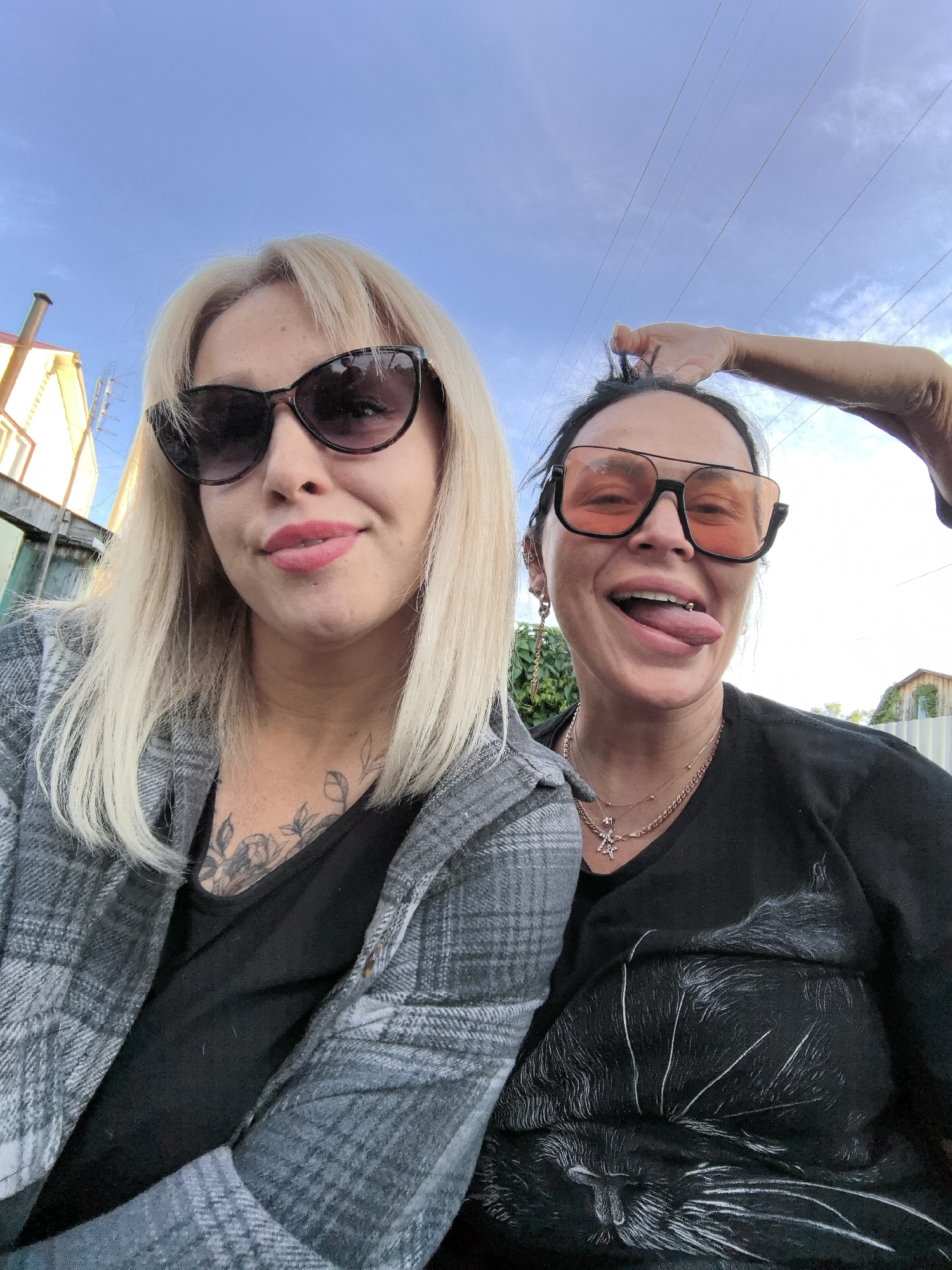 Two women are captured in a close-up selfie outdoors, likely in a residential area of Noviy, Russia, under a clear blue sky. The overall mood is casual and playful.
On the left, a woman with blonde hair and dark, cat-eye sunglasses is visible from the chest up. She wears a grey plaid open shirt over a black top, revealing tattoos on her upper chest and neck. Her lips are painted red, and she has a subtle smile, looking directly at the camera. Reflections in her sunglasses show distorted elements of the surroundings, possibly the person taking the photo or parts of a building.
To her right, partially obscured by the first woman, is another woman with dark hair, wearing large, orange-tinted sunglasses. She has a lively expression, sticking out her tongue, and one of her hands is raised to pull up her hair from the top of her head. She wears a black t-shirt featuring a detailed white illustration of a cat's face and fur. Several necklaces, including one with a cross pendant, adorn her neck. She also has multiple ear piercings and a tongue piercing visible.
The background features residential elements. On the far left, part of a light-colored building with a chimney is visible. Towards the right, beyond the women, more faint architectural structures and a wooden fence can be seen, suggesting a backyard or a street with houses. Overhead, against the bright blue sky, several thin power lines stretch across the frame. The lighting suggests it is daytime, possibly late afternoon due to the clear, bright sky. There is no discernible text visible in the image.