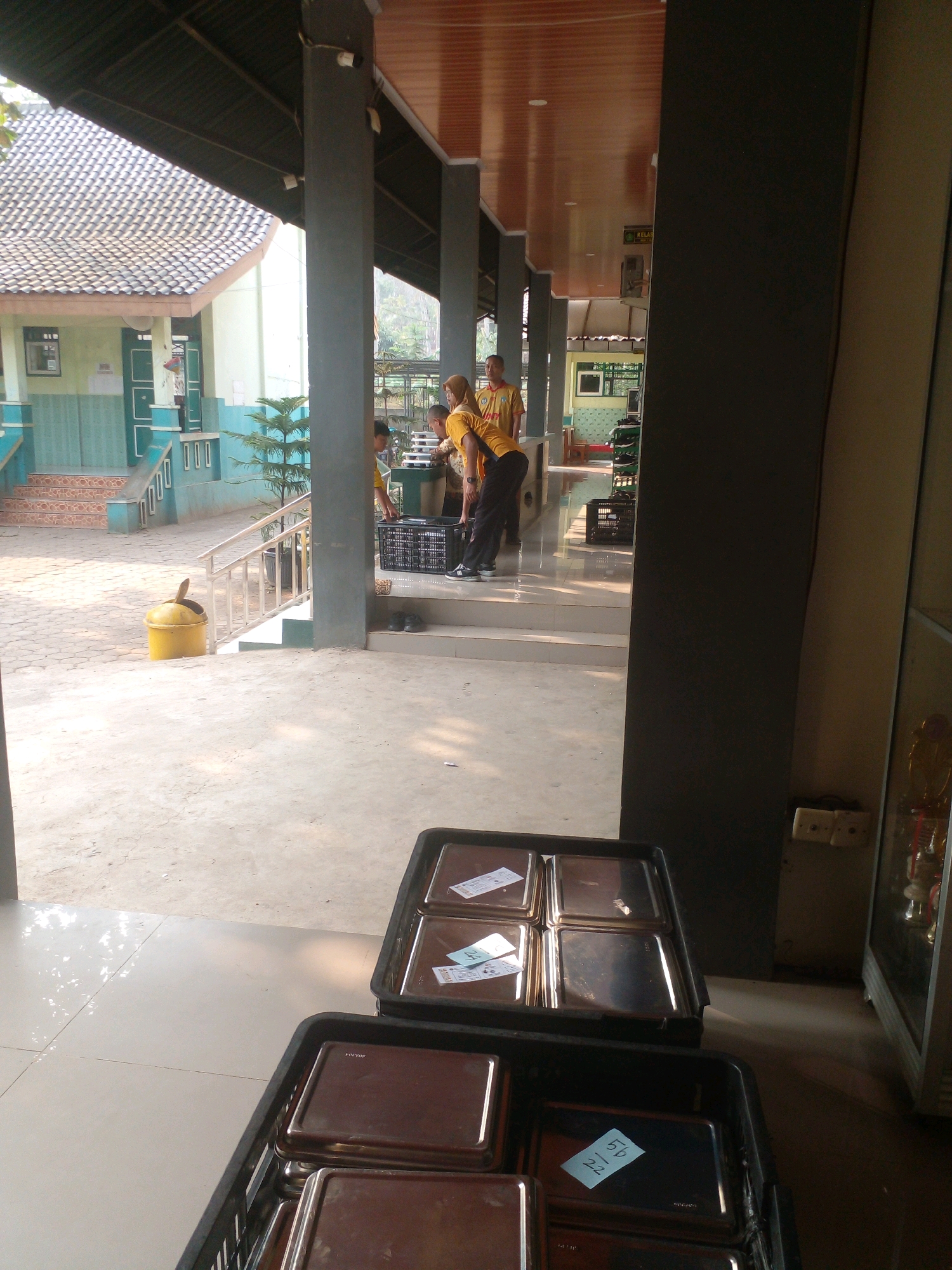 The image depicts an outdoor scene in front of a building, possibly a school or public facility, located in Banyuputih, Indonesia. In the foreground, two black plastic crates are filled with metal trays, some of which have labels. The crates are placed on a tiled floor. In the mid-ground, several individuals, appearing to be students or staff wearing yellow shirts, are engaged in activity. One person is carrying a tray, while others are standing near a black plastic crate. To the left, a yellow trash can is situated near a set of stairs with a white railing. The background shows a building with a tiled roof, a staircase, and trees. The environment appears to be sunny.
