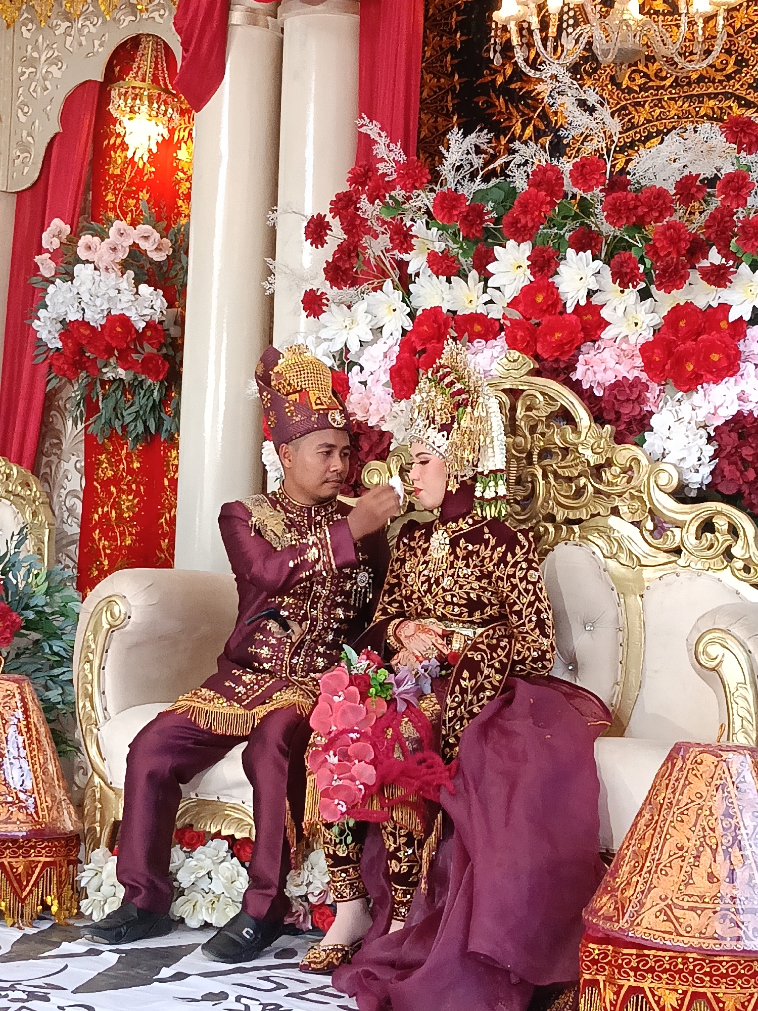 This image captures a wedding ceremony in Geulumpangsulu Timu, Indonesia. A bride and groom are seated on a white and gold ornate couch, dressed in traditional maroon and gold attire adorned with intricate patterns. The groom wears a matching hat, while the bride sports a tall, elaborate headdress with gold accents. The groom is gently feeding something to the bride. The backdrop is richly decorated with abundant red, white, and pink flowers, along with draped red fabric and an ornate chandelier. Flanking the couch are decorative lamps with intricate gold patterns. The setting suggests a formal and festive occasion.