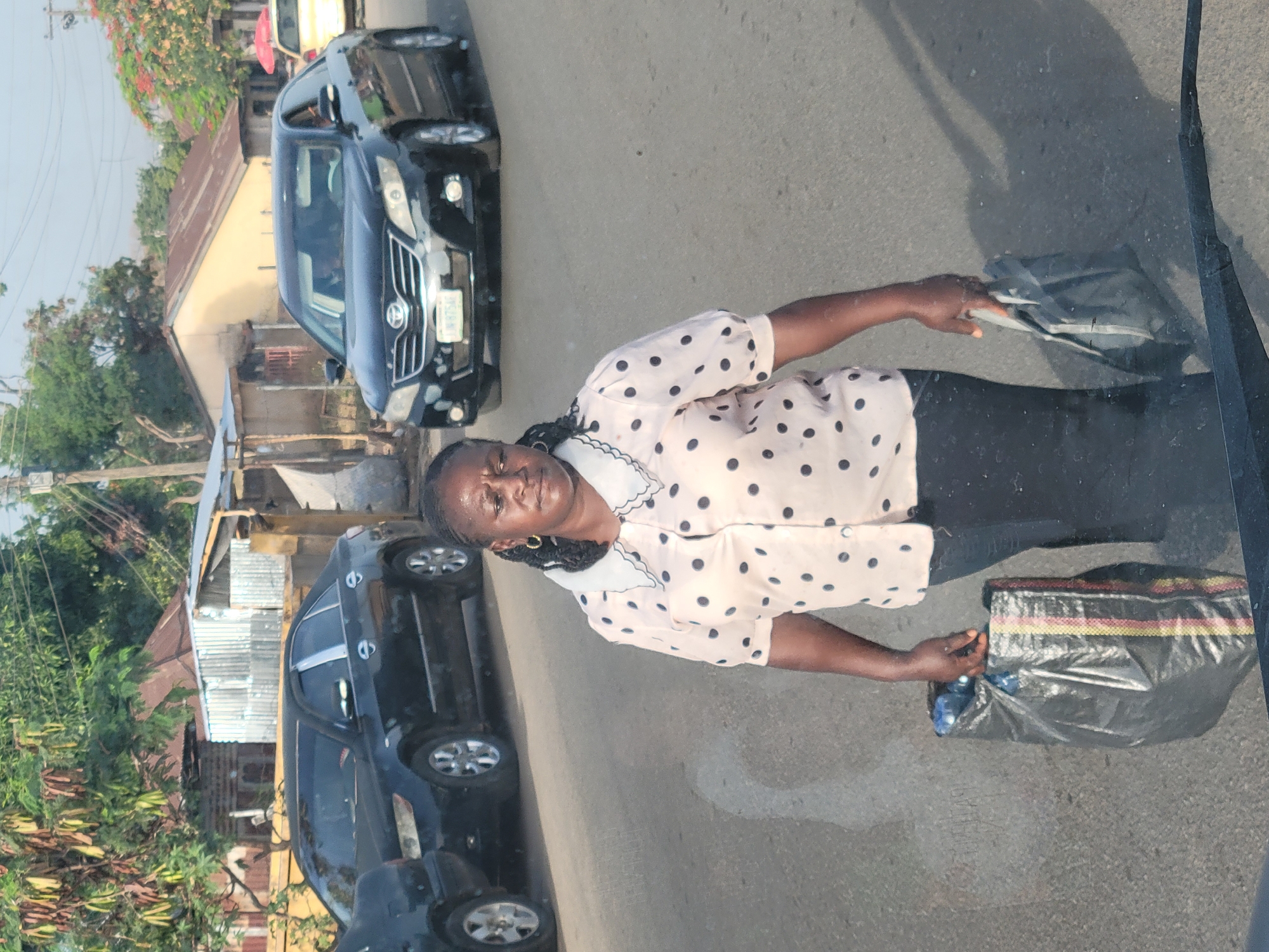 The image depicts an adult woman standing on an asphalt road, positioned between parked cars and a backdrop of buildings and greenery, under bright daylight.
The central subject is a woman of African descent with dark skin and braided hair. She is wearing a light-colored, short-sleeved shirt with dark polka dots and a white collar, paired with dark trousers. She holds two bags: a lighter-colored bag in her right hand and a dark plastic bag, possibly striped with red, yellow, and green, in her left hand. The dark bag appears to contain a blue bottle, possibly water. Her facial expression is neutral.
Behind the woman, two black cars, likely SUVs or sedans, are visible on the road. The front license plate of the closer car is partially legible, displaying characters that resemble "N8754E," indicative of a Nigerian registration.
The background features several buildings with corrugated metal roofs and lush green trees and bushes, suggesting a semi-urban or residential environment. The scene is bathed in clear, bright sunlight, casting distinct shadows on the ground, indicating daytime. The woman appears to be standing, possibly waiting or walking, along the street. A reflection, likely from the window of the vehicle from which the photo was taken, is noticeable on the bottom right of the image. The setting is consistent with a street scene in Jos, Nigeria.