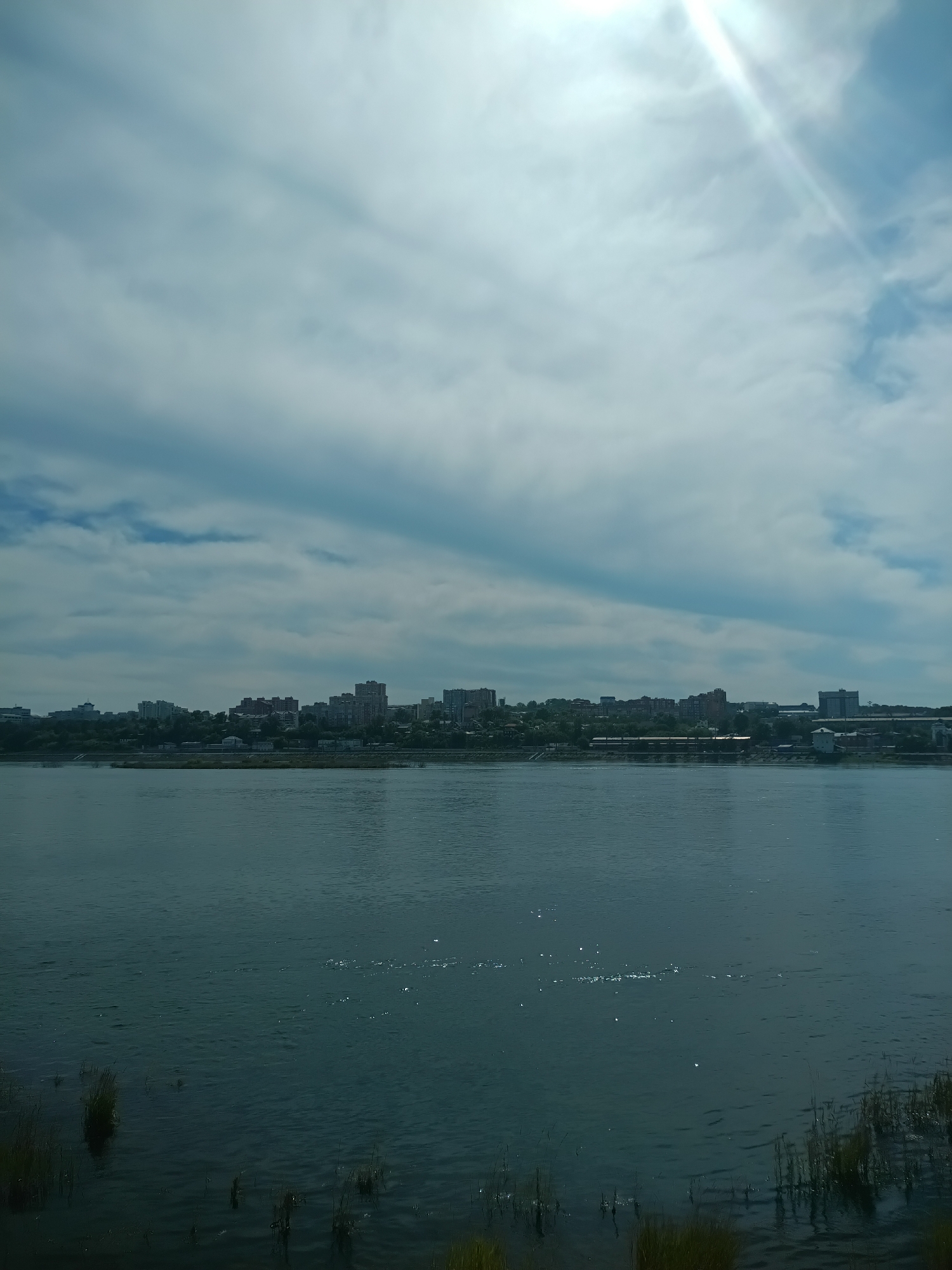 The image depicts a wide, dark blue river flowing in the foreground. The water's surface is rippled, with small white glints of light suggesting it is reflecting sunlight. Tall, dark green reeds are visible along the riverbank on the lower right side.
In the background, across the river, is a city skyline with numerous multi-story buildings of varying heights and architectural styles. The buildings are predominantly in shades of grey, brown, and white. A line of trees separates the city from the river.
The sky above is a mix of light blue and white, with several large, fluffy clouds. A bright sunbeam, partially obscured by clouds, shines down from the upper right corner of the frame, creating a lens flare effect. The overall impression is a bright, partly cloudy day over a cityscape by a river. This scene is characteristic of the Irkutsk embankment along the Angara River in Irkutsk, Russia, which separates the historical center from the newer districts.