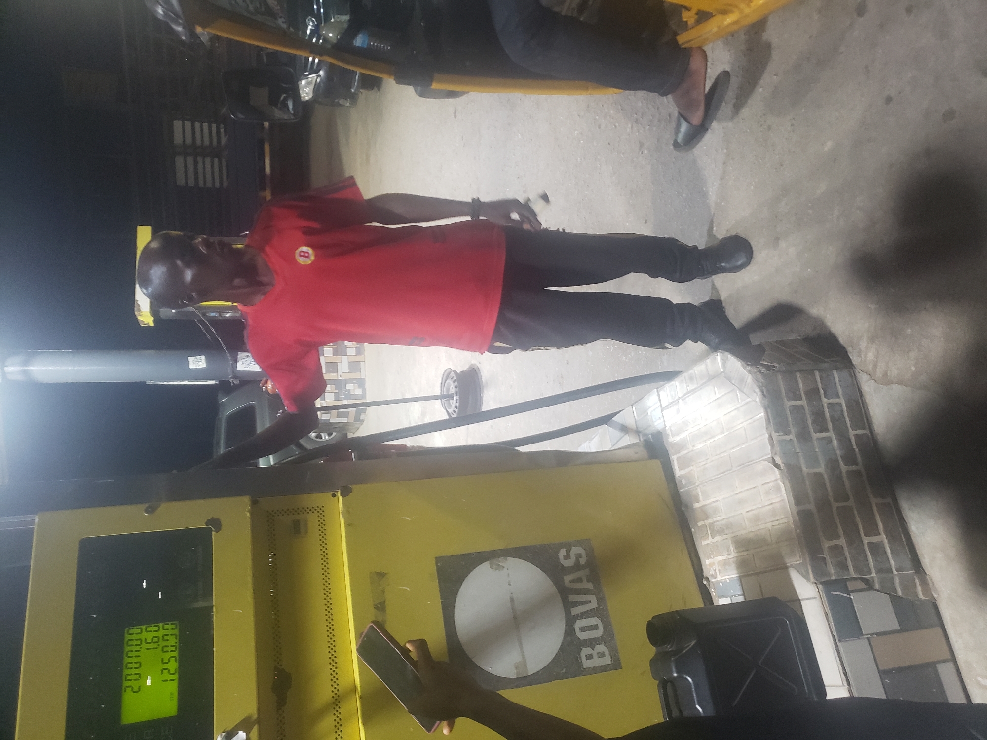 An individual in a red shirt is standing at a yellow gas pump with the logo "BOVAS." The pump indicates a value of "200000.00" and a price of "12500.91." Another individual is holding a smartphone near the base of the pump. A jerrycan sits near the pump's base. The location is in Alalubosa, Nigeria.