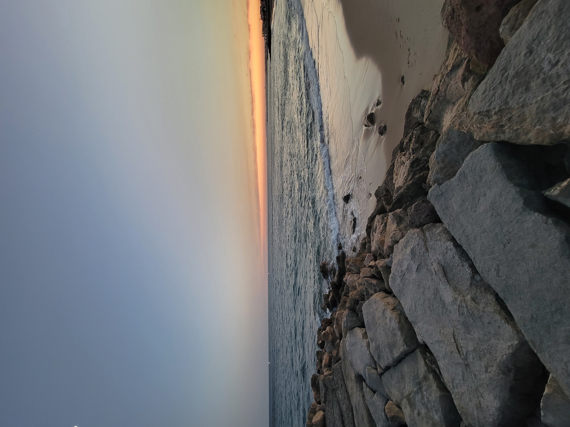 The image depicts a sunset scene at the beach in Quarteira, Portugal. In the foreground, large, rough-hewn grey and brown rocks form a breakwater or seawall. Beyond the rocks, a sandy beach meets the gentle waves of the ocean. The water is a deep blue-green, with small whitecaps indicating the tide. In the distance, the horizon is illuminated by the warm hues of the setting sun, with shades of orange and yellow bleeding into the pale blue sky. A sliver of the moon is visible in the upper left quadrant of the sky. The coastline in the far distance features a few scattered lights, suggesting a town or settlement. The scene is tranquil and natural, with no people or animals visible. The overall impression is one of peaceful solitude at the end of the day.