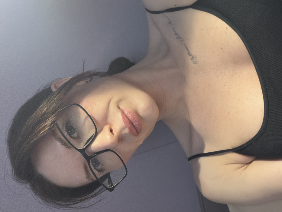 The image displays a close-up, angled portrait of a female subject, estimated to be between 20 and 35 years old, in a domestic setting within the city of Perm, Russia. The subject has dark brown hair, pulled back from her face, and light-colored eyes. She is wearing black-framed rectangular eyeglasses. Her lips are a light pink, appearing glossy. A tattoo of handwritten script is visible on her upper right chest, near the collarbone and extending towards the shoulder. She is dressed in a black, ribbed spaghetti-strap top, exposing her shoulders and upper chest, which exhibit visible freckles. The subject is illuminated by a distinct light source from the upper left, creating highlights on her face and a shadow across her left cheek. The background is an out-of-focus, solid, light purple wall. The subject is looking directly at the camera, with a slight, neutral expression.