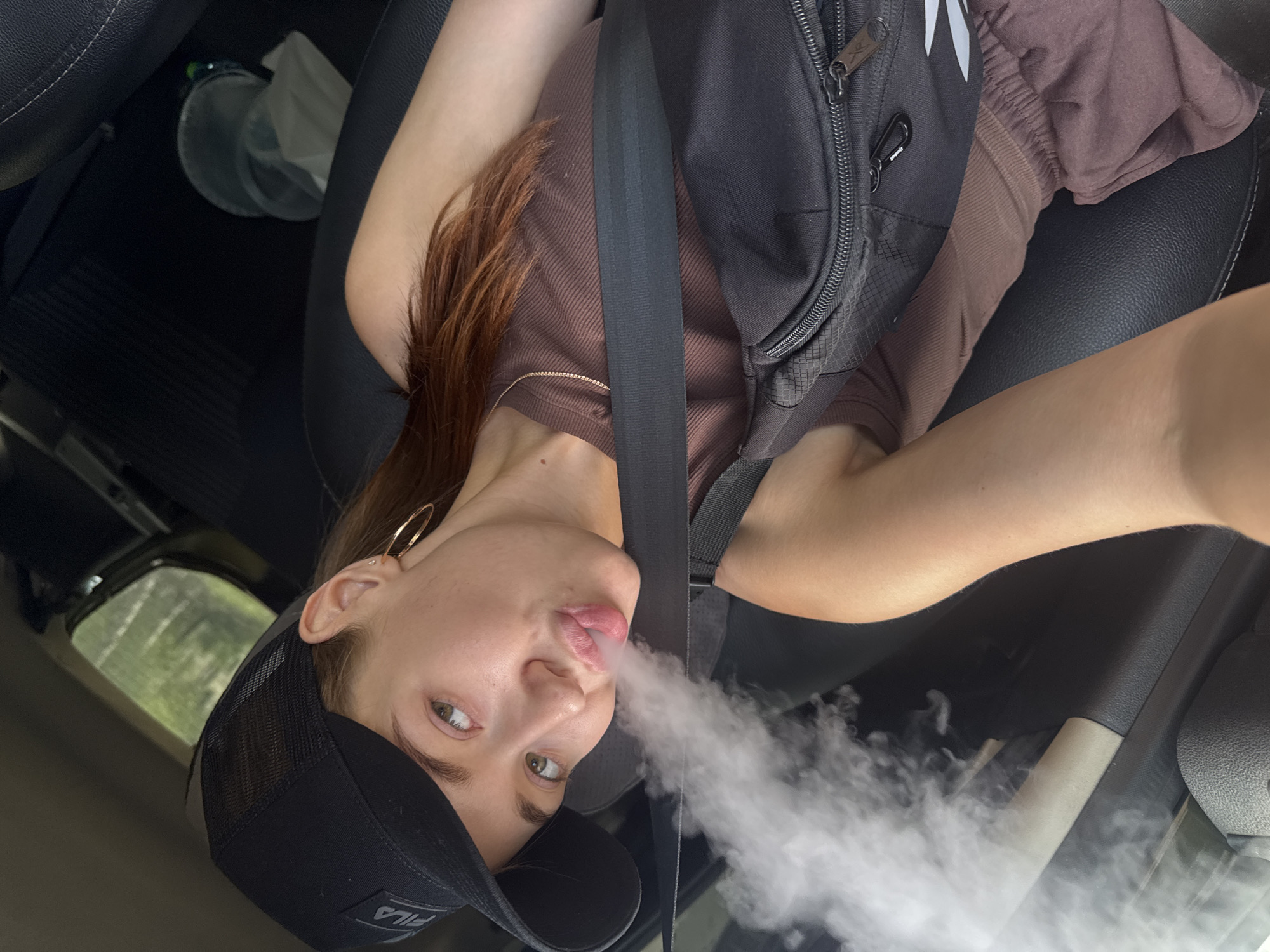 A young woman with auburn hair is visible in the driver's seat of a car, exhaling a cloud of vapor. She is wearing a brown top, a black fanny pack, and a black baseball cap. A gold hoop earring adorns her left ear. The car's interior, including a seatbelt and a black dashboard, is visible. Outside the car window, trees and foliage suggest a natural, outdoor setting. The lighting indicates it is daytime. There is no visible text in the image. The activity suggests the woman is vaping.