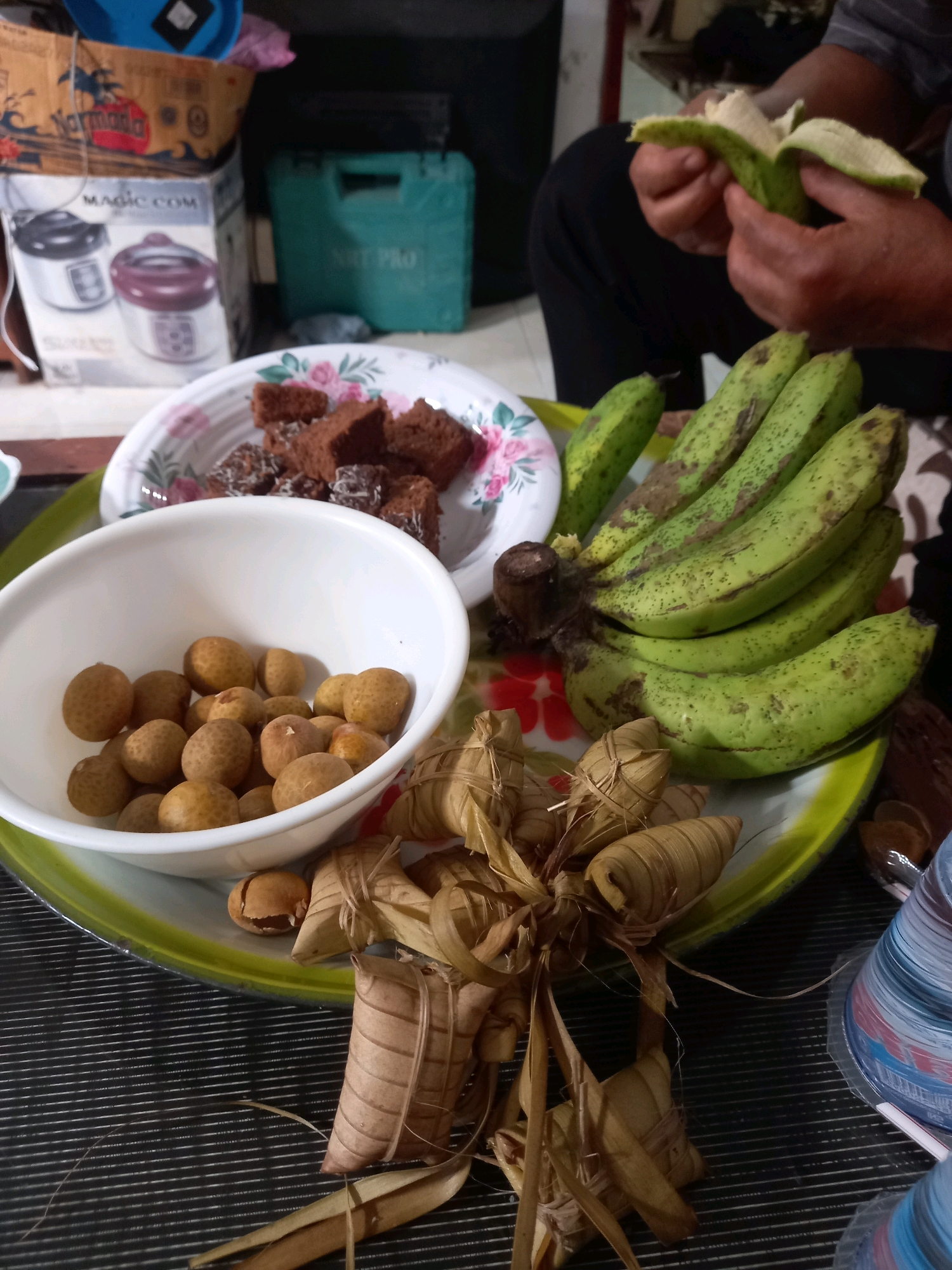 The image depicts a close-up view of a spread of food items, likely prepared for a gathering or meal. The setting appears to be indoors, with a table surface covered in a striped mat. In the foreground, a cluster of several wrapped food items, possibly rice cakes or desserts, bound with dried leaves and string, are arranged on a yellow and green tray. To the left of these wrapped items, a white bowl is filled with small, round, brown fruits, resembling longans. Behind the bowl of fruit, a white plate with a floral rim contains several pieces of dark brown, rectangular confections, appearing to be brownies or a similar cake. A bunch of unripe, green bananas, some with brown speckles, are also present on the tray, positioned behind the plate of confections. In the upper right of the image, a person's hands are visible, peeling a green banana. In the background, shelves are stocked with various items, including boxes labeled "Normada" and "MAGIC COM," and a green plastic container. The presence of a person peeling a banana suggests the food is being prepared or served. The overall scene is one of casual food presentation, possibly in a residential setting in Mataram, Indonesia, given the types of food items visible and the general aesthetic.