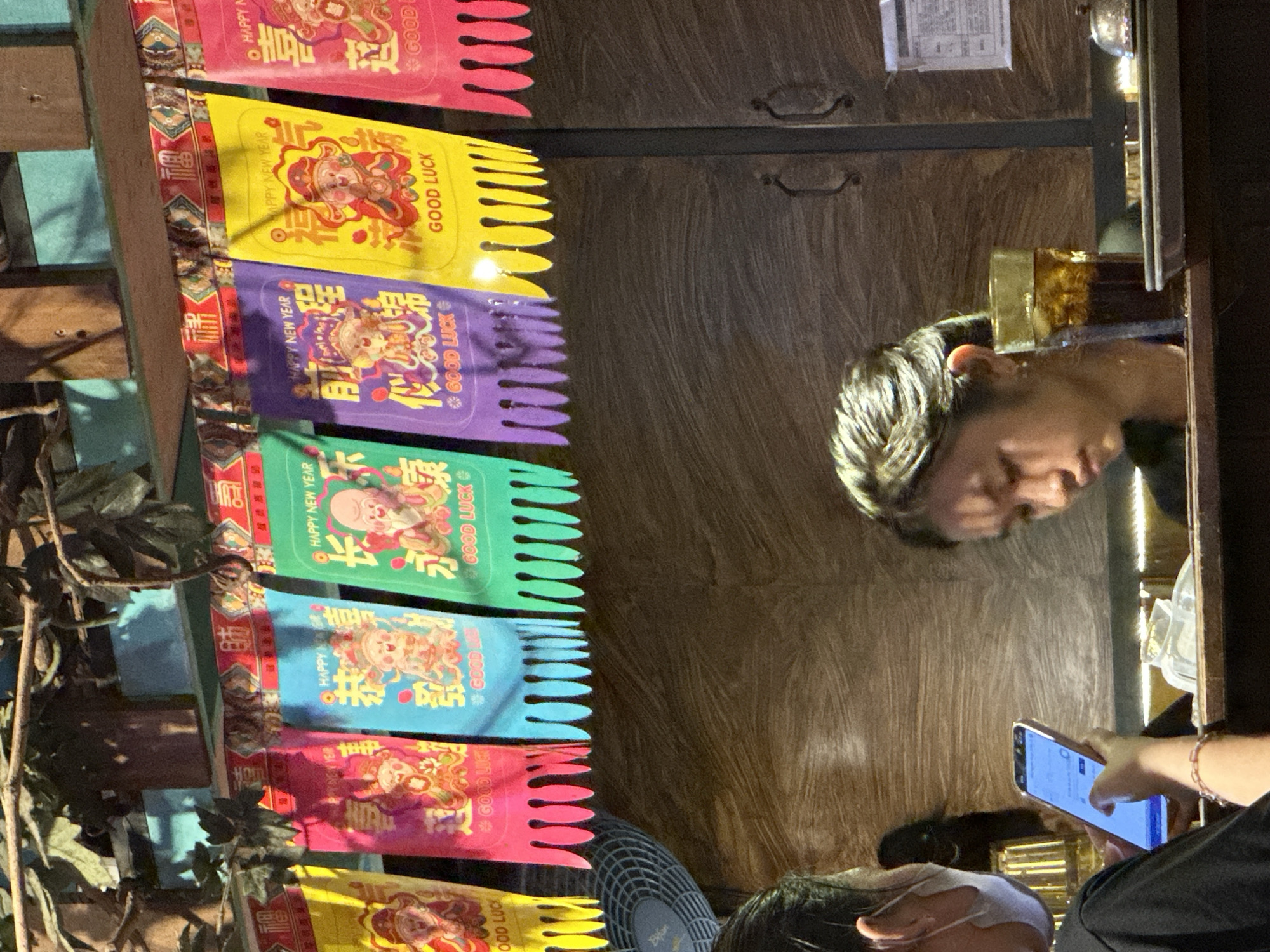 In this indoor shot, a person is visible from the shoulders up, looking downwards with a contemplative expression. They are holding a smartphone in their right hand, interacting with it. The background is a textured wooden wall, with a dark wooden cabinet and a glass pane. A row of colorful packets, likely red envelopes or good luck charms, are strung vertically along the left side of the frame. These packets feature Chinese characters and images of figures, with text such as "HAPPY NEW YEAR" and "GOOD LUCK" visible. The lighting suggests it could be evening or an indoor environment with artificial light. The scene has a somewhat casual and possibly festive atmosphere due to the decorative packets. The inclusion of a smartphone and the casual attire suggest a modern, everyday setting. The presence of "HAPPY NEW YEAR" text hints at a recent or upcoming Lunar New Year celebration.