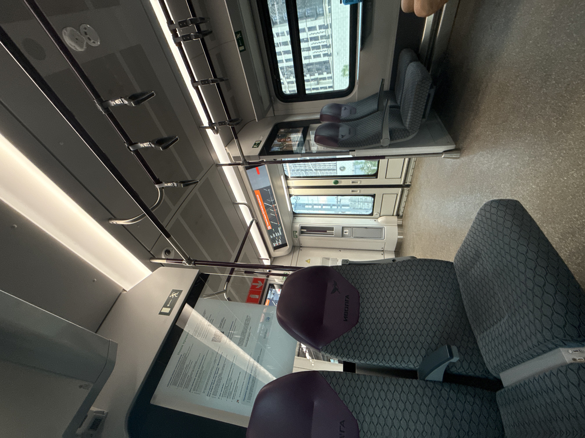 The image shows the interior of an "Ivolga" train, presumably somewhere in an urban area. The seats are patterned and appear to be made of cloth. There are two sets of seats visible in the foreground, and another set in the background. The train has windows showing the outside, with buildings visible beyond the glass. A display screen on the interior has some text. The image is taken at an angle.