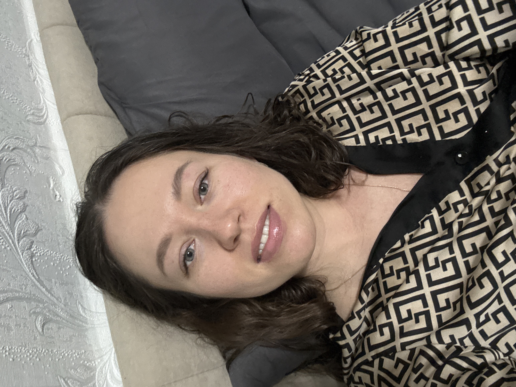 The image shows a woman lying down, facing the camera, with her head resting on a textured, off-white surface, likely a sofa or bed. A dark grey pillow is visible behind her head. She has fair skin, brown curly hair, and light-colored eyes. She is smiling slightly, and her lips appear glossy. She is wearing a top with a geometric pattern in beige and black, with a black collar. The background consists of a textured white surface, possibly wallpaper, on the left. There is no explicit indication of the location in the city of Ферма, Russia, or any actions or interactions taking place. The scene appears to be indoors.