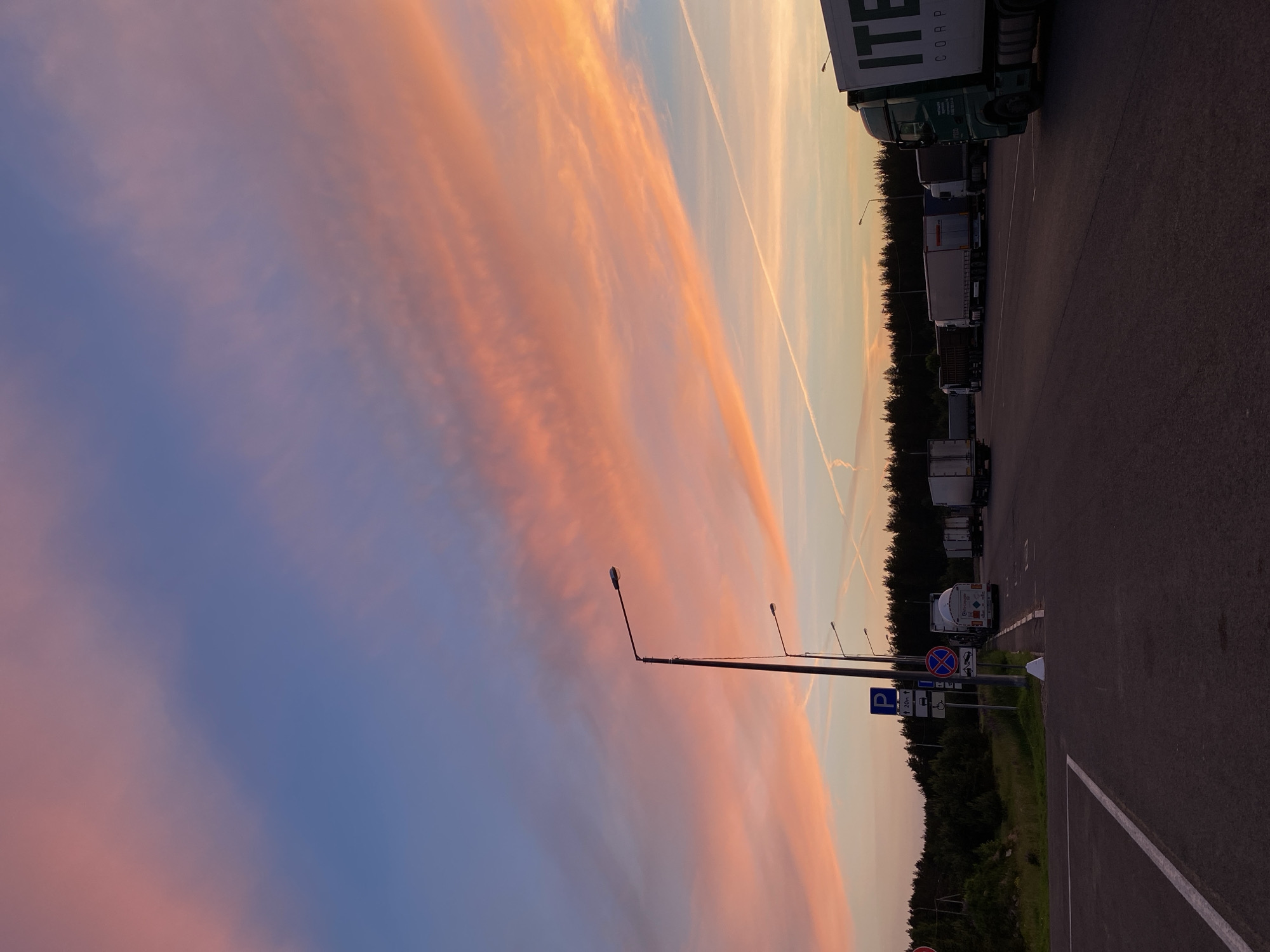 A wide-angle, low-angle shot captures a truck stop with a dramatic sunset sky over Сазоново, Russia. The sky is a canvas of blues, oranges, and pinks, with wispy clouds and contrails streaking across the horizon. In the foreground, a paved asphalt surface with white lane markings leads into the frame. A row of parked semi-trailer trucks is visible, stretching into the distance. One truck in the foreground, on the right side of the image, is green and white, with the branding "ITE CORP" prominently displayed. Streetlights stand tall along the edge of the parking area, casting a warm glow. Several traffic signs are present near the entrance to the truck stop, including a blue parking sign and a red circular sign with a diagonal line. A dense line of dark green trees forms the backdrop, silhouetted against the colorful sky. The scene appears to be during twilight, just after sunset. There are no people visible in the image.