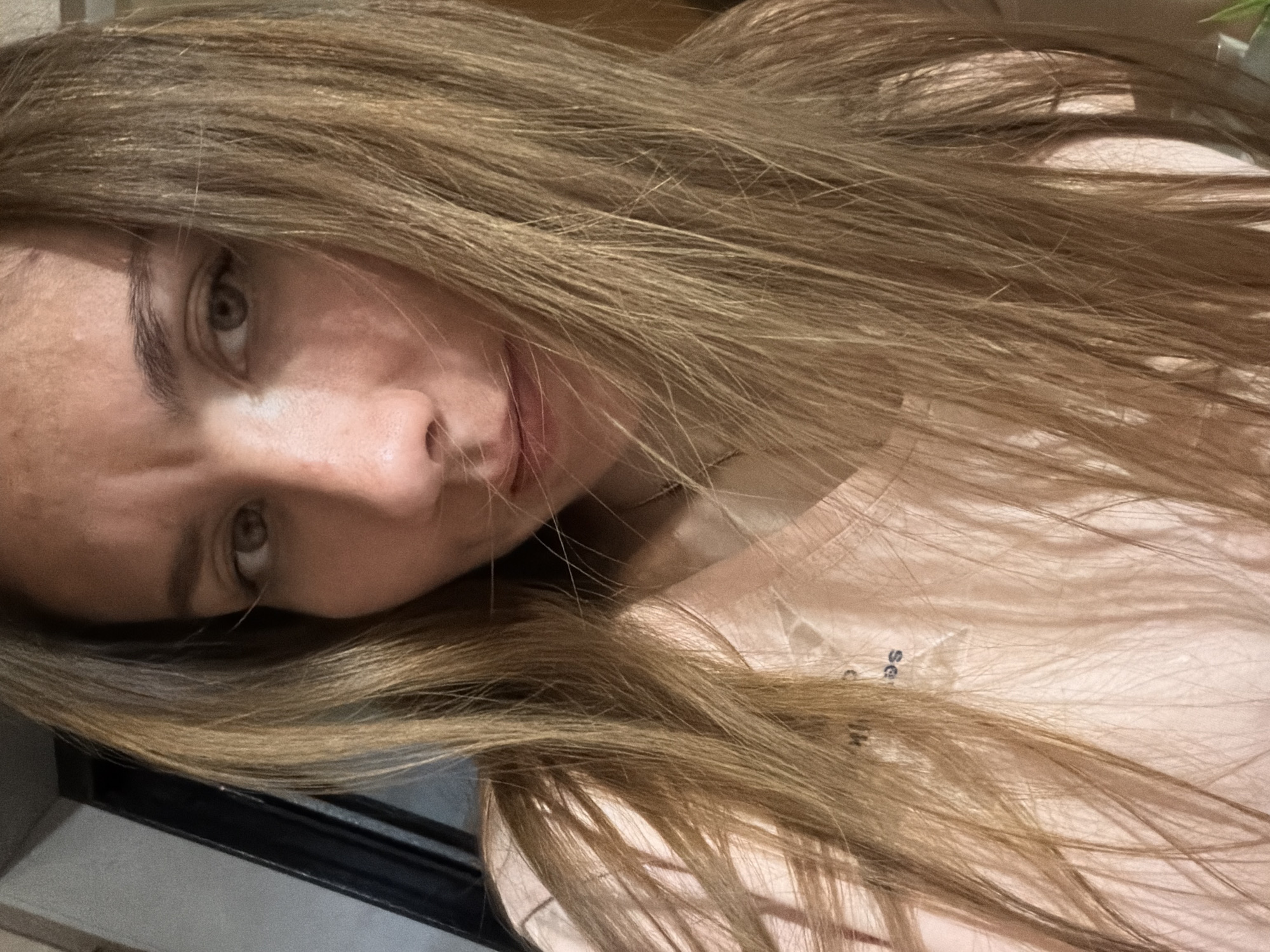 A close-up, selfie-style photograph captures a young woman's face from the chest up. She has long, wavy, light brown hair that frames her face and falls across her shoulders and torso. Her eyes are a hazel color and she is looking directly at the camera. Her eyebrows are slightly furrowed, giving her a concerned or questioning expression. Her nose is visible, and her lips are slightly parted. She is wearing a light pink t-shirt, and a faint logo or text is visible on the fabric. The background is blurred and appears to be an indoor setting, with a dark vertical element on the left and a lighter wall on the right. There is no indication of the city of Tambov, Russia, in the image.