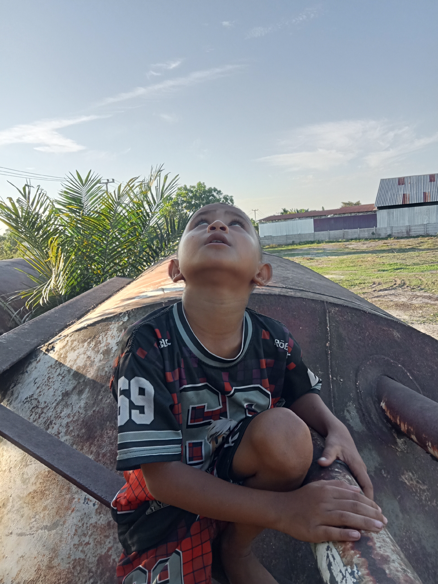 A young boy with dark skin and short black hair is crouching on a large, weathered, cylindrical metal object. He is looking upwards, with his mouth slightly open and his eyes wide. He is wearing a black t-shirt with colorful geometric patterns in red, white, and gray, and the number "69" is visible on the left sleeve. His shorts are black with a red diamond pattern. His hands are resting on his knees and the metal surface.
The metal object appears to be industrial or a large piece of discarded equipment, with rust and discoloration visible on its surface. Behind the boy, palm fronds and trees are visible, and further in the background, there are buildings with corrugated metal roofs and a fence. The ground is a mix of grass and dirt. The sky above is a clear blue with some wispy clouds. The lighting suggests it is daytime, likely late afternoon due to the shadows.
The location is identified as Kualu, Indonesia. This appears to be an outdoor scene, possibly an industrial area or a rural setting with some infrastructure.