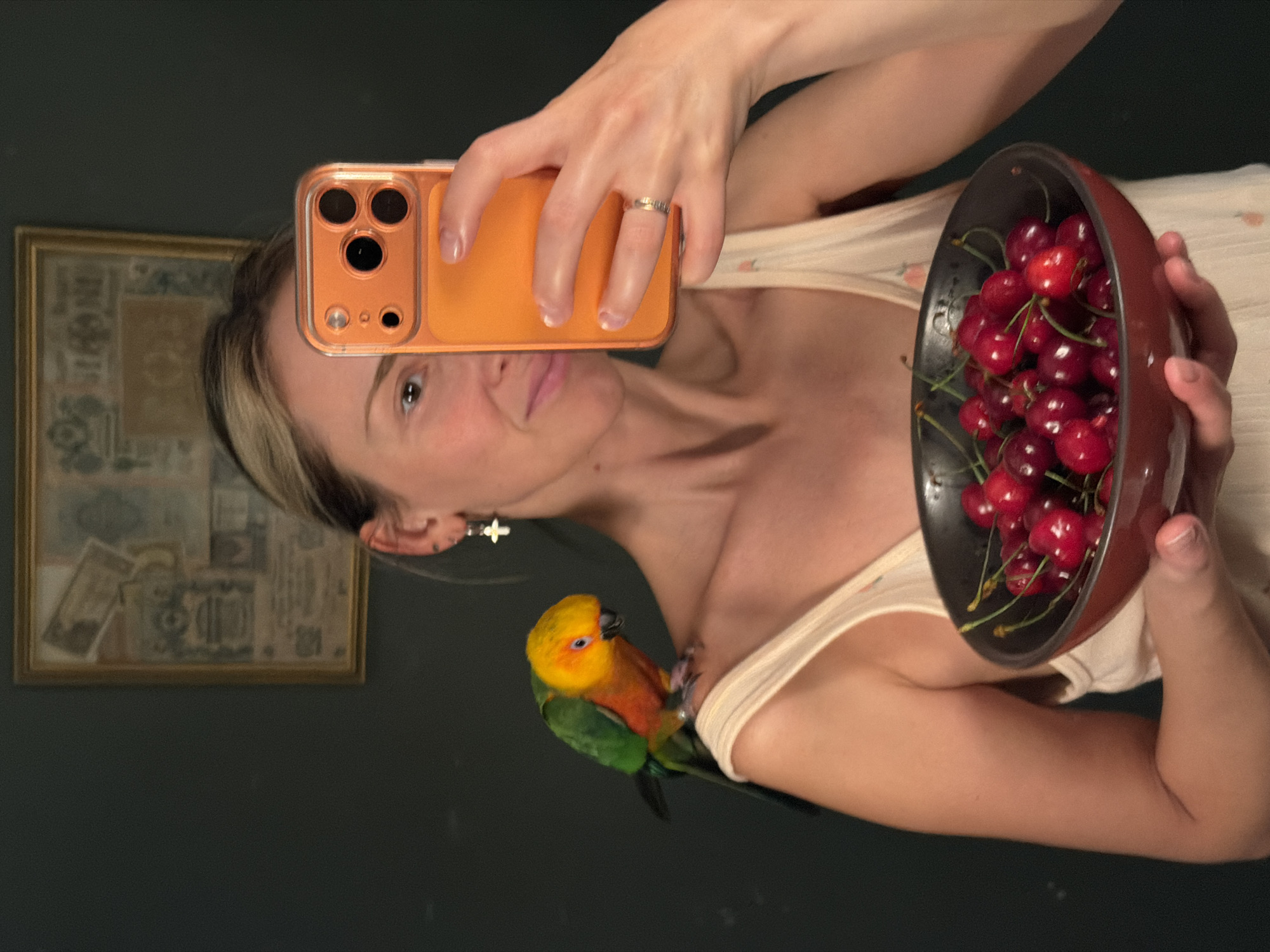A selfie taken indoors, possibly in a home, shows a woman holding an orange-cased smartphone in her right hand and a bowl filled with red cherries in her left hand. A colorful parrot is perched on her left shoulder. She wears a light-colored tank top and a ring on her right hand. Behind her is a framed piece of art on a dark green wall. There is no visual information in the image to connect the scene with the city of Khimki, Russia.