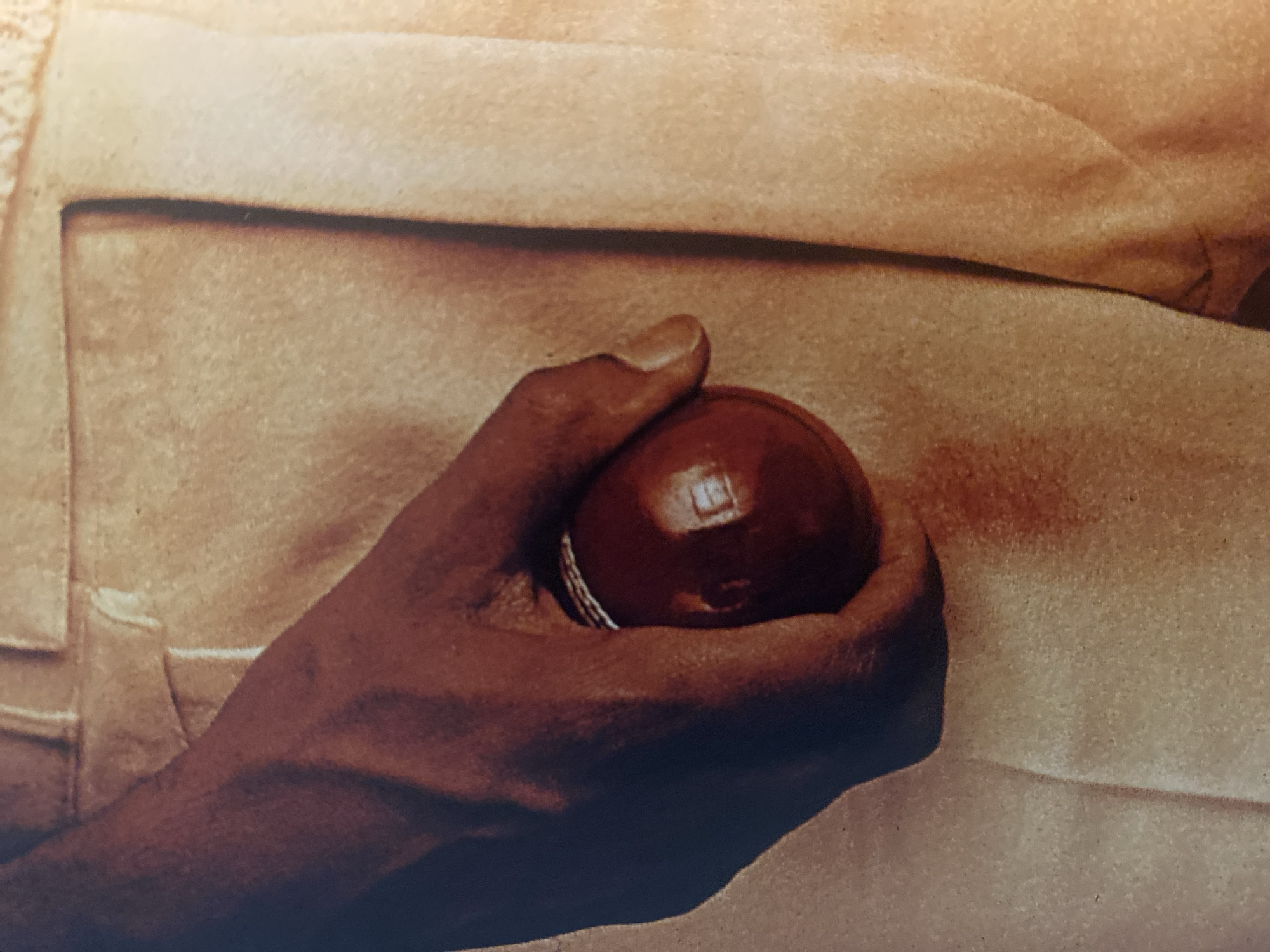 A close-up depicts a brown-skinned hand holding a red spherical object. The hand is positioned against a light tan background that appears to be textured fabric, possibly clothing. A pocket seam is visible on the left side of the frame. The location in the city of Шебунино, Russia is not discernible from the image.