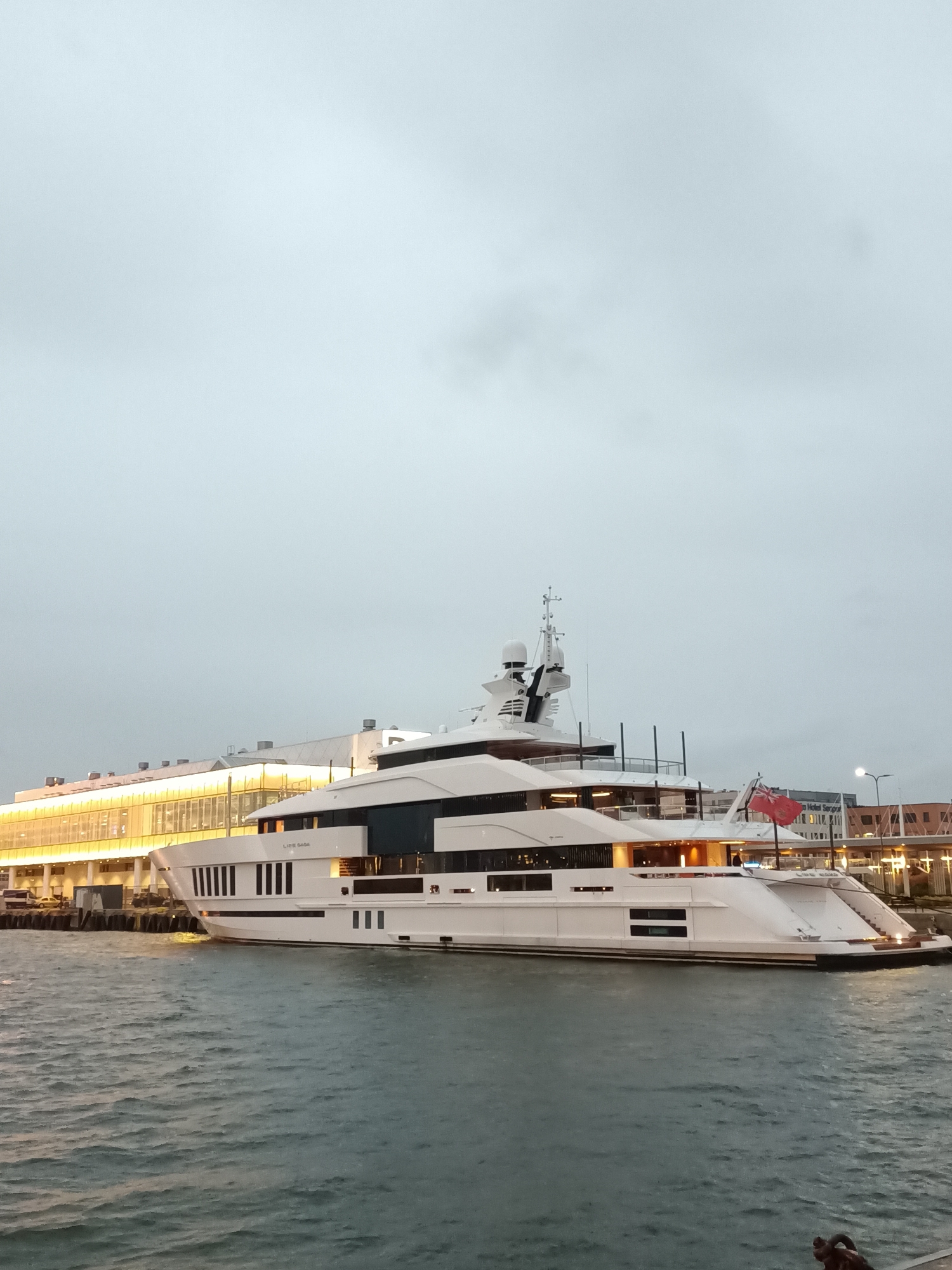 A large, white luxury yacht, named "LIFE SAGA", is docked at the pier in Tallinn, Estonia. The yacht is the primary focus of the image, with its sleek design and multiple decks clearly visible. A red flag flies from the stern of the yacht. Behind the yacht, a modern building with large windows, illuminated from within, is visible. The sky is overcast, and the water is choppy. The overall scene suggests a calm, perhaps evening or early morning, setting in a maritime area of the city.