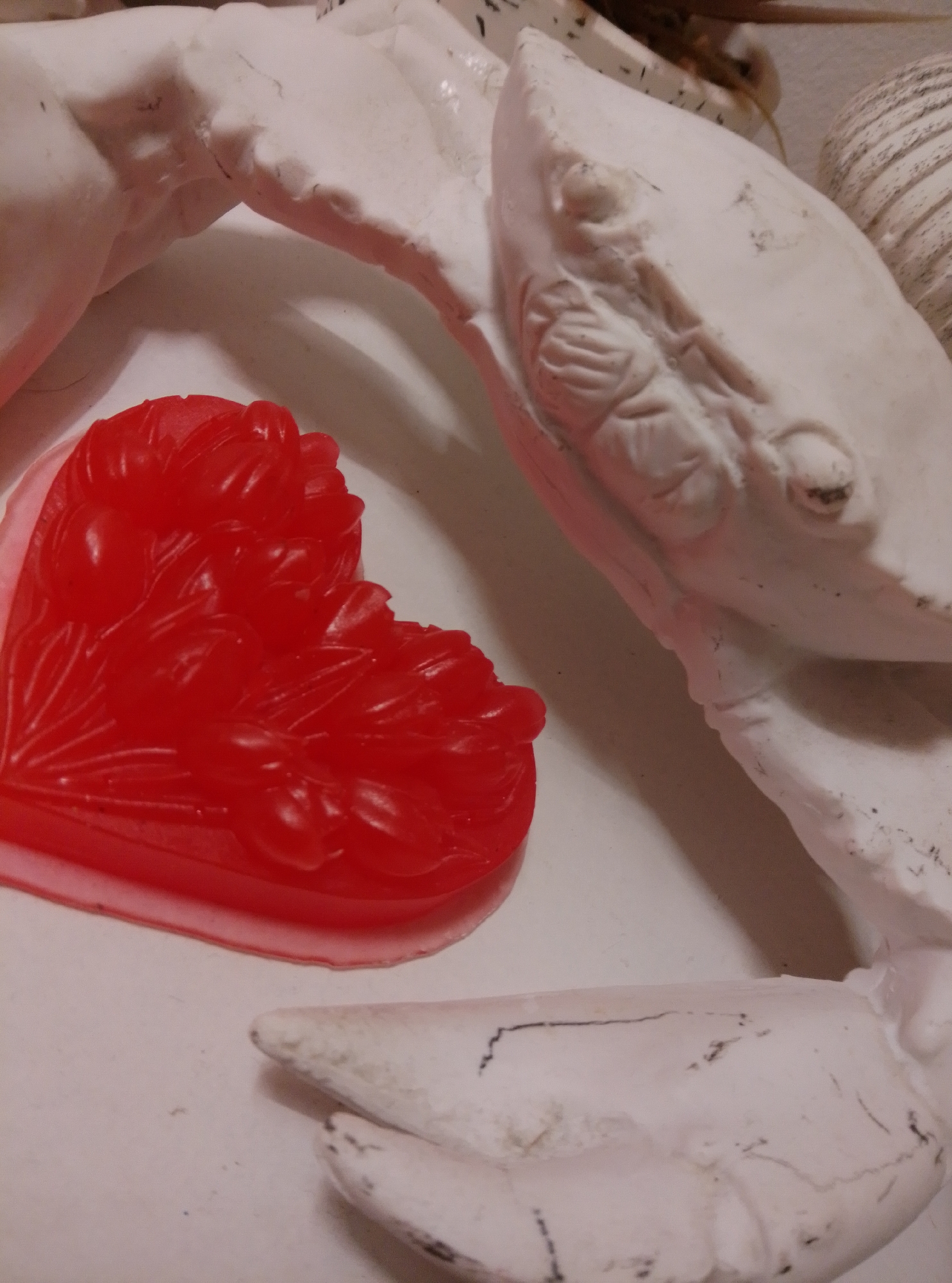 A red, heart-shaped object with a raised floral design rests on a white surface. Behind it, several abstract white forms are visible, some appearing to be parts of a larger sculpture. The lighting is soft, casting subtle shadows. The scene is not specific to Wedel, Germany.
