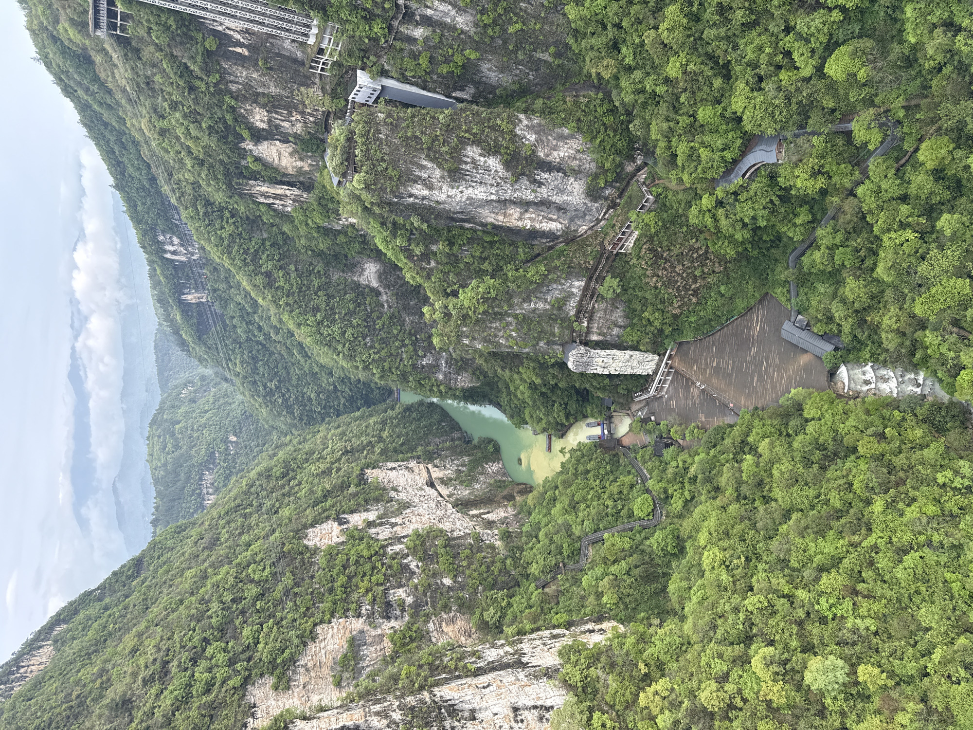 A high-angle shot shows a mountainous, heavily forested landscape in Zhangjiajie, China, featuring gray rock formations, green foliage, and a river winding through the valley. A large dam structure is present on the right side of the frame. Man-made features include walkways, a building on a cliff, and a cable car structure extending down the mountain on the left. The sky is overcast, adding to the subdued light of the scene.