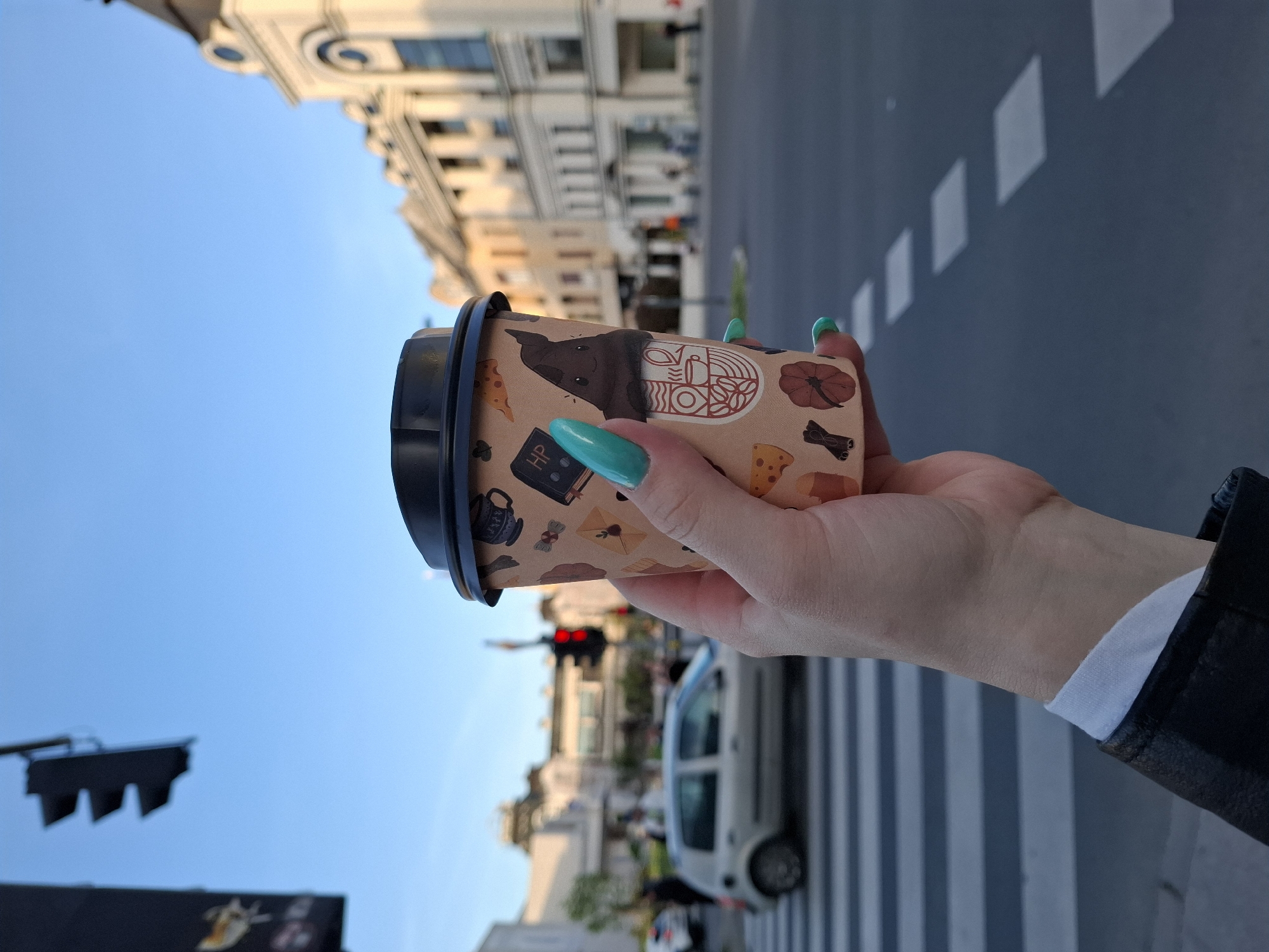 The image shows a person holding a coffee cup with a black lid in their right hand. The cup has a light brown background with various cartoon-like images on it. The person has long, bright teal-colored fingernails. In the background, there is a city street with a pedestrian crosswalk. A car is partially visible, and a traffic light is showing a red light. Buildings can be seen on both sides of the street. The sky is blue. The location is Unknown.