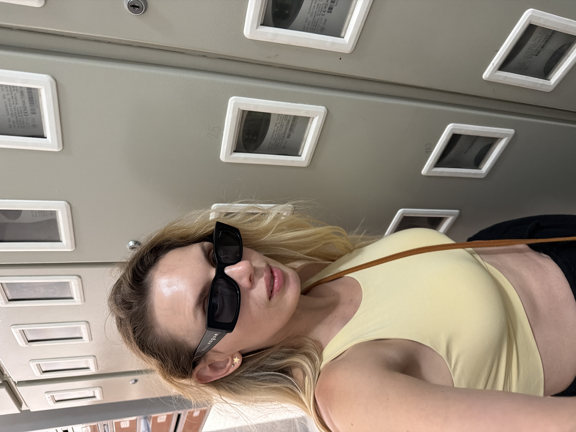 A woman with blonde hair and sunglasses is posing for a selfie in front of a row of gray metal cabinets. She is wearing a yellow tank top and black pants. The cabinets appear to be utility meters, with small windows showing dials and readings. One of the cabinets has the number "25" written on it. The overall environment suggests an indoor urban setting, possibly a public building or facility. The background includes more of the cabinets and some visible office supplies on a shelf to the left. The lighting is bright and natural, indicating daytime. The location is identified as Budva, Montenegro.