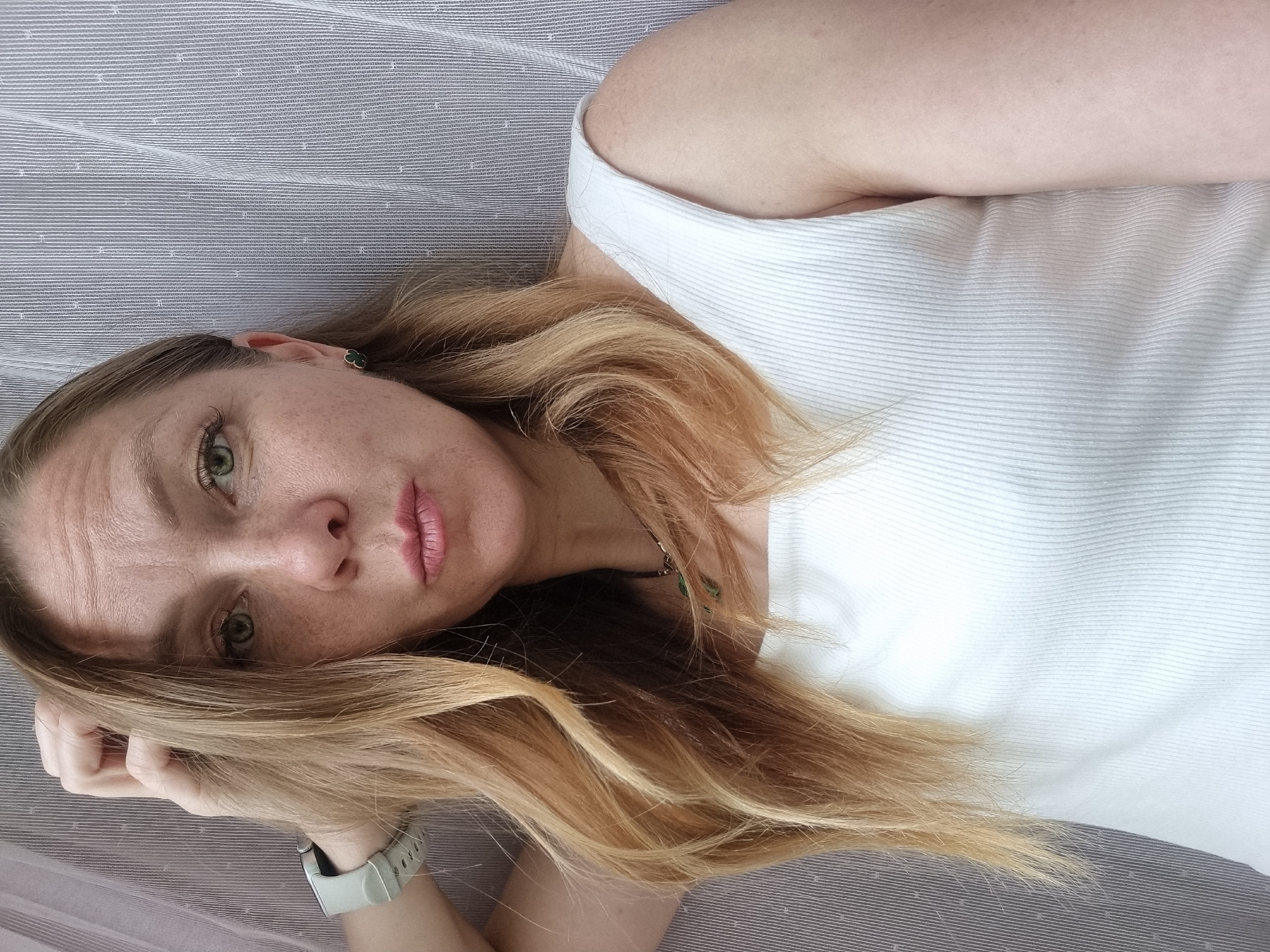 A woman with blonde hair and green eyes is looking at the camera, her left hand touching her hair. She is wearing a white tank top and a necklace. Her left arm is visible, resting on her head, and she has a watch on her left wrist. She is indoors, in front of a gray and white striped curtain.