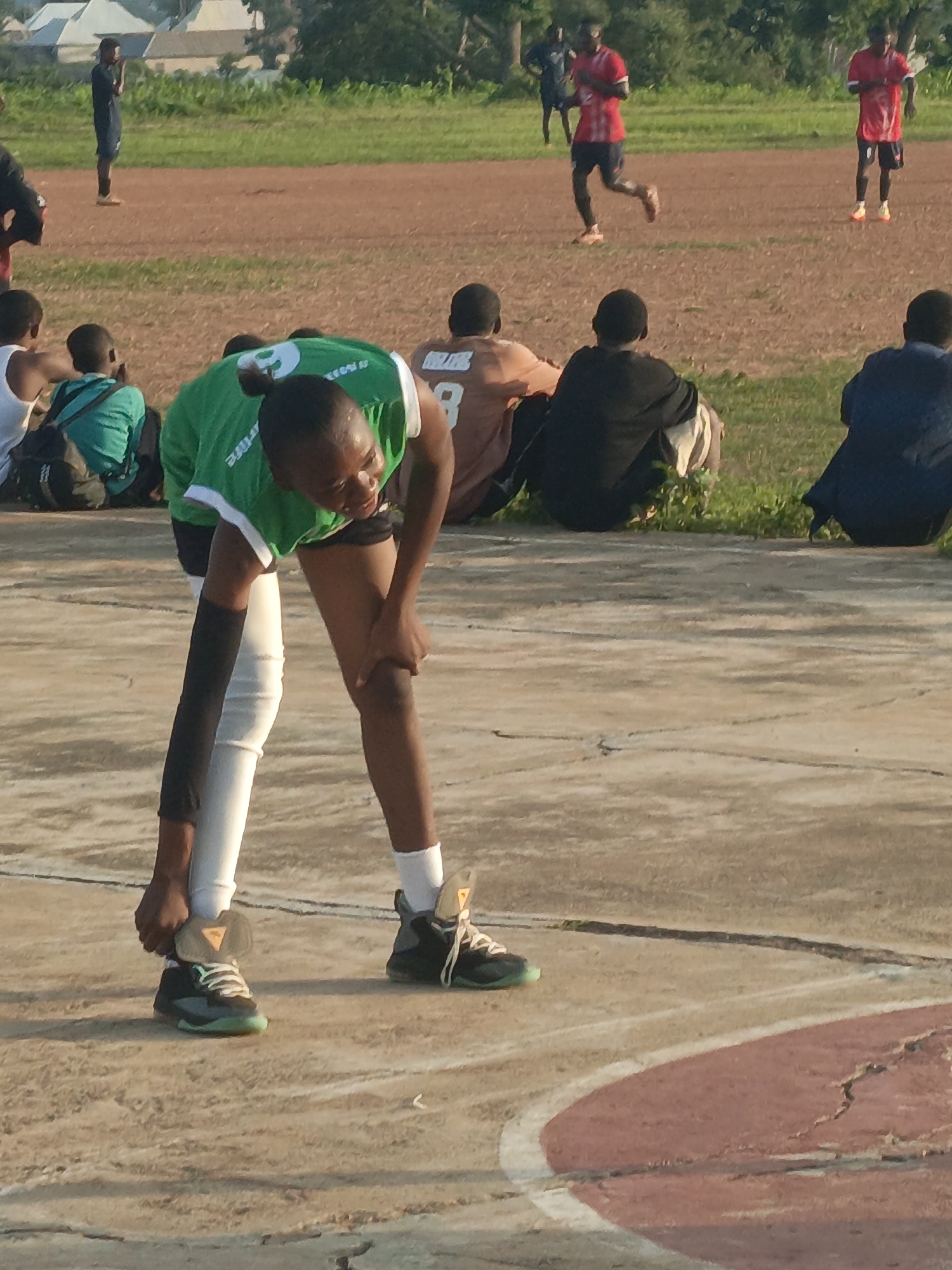 A group of young people, mostly male, are gathered on an outdoor sports field in Akwanga, Nigeria. The foreground is dominated by a young person wearing a green shirt and white leggings, bending down to adjust their shoelaces. They are wearing black and teal athletic shoes. Behind this person, several other individuals are seated or standing, some watching the activity. In the background, more young men in red shirts are playing soccer on a dirt field. The environment appears to be a sports complex with a running track and open grassy areas. The lighting suggests it is daytime, possibly late afternoon.