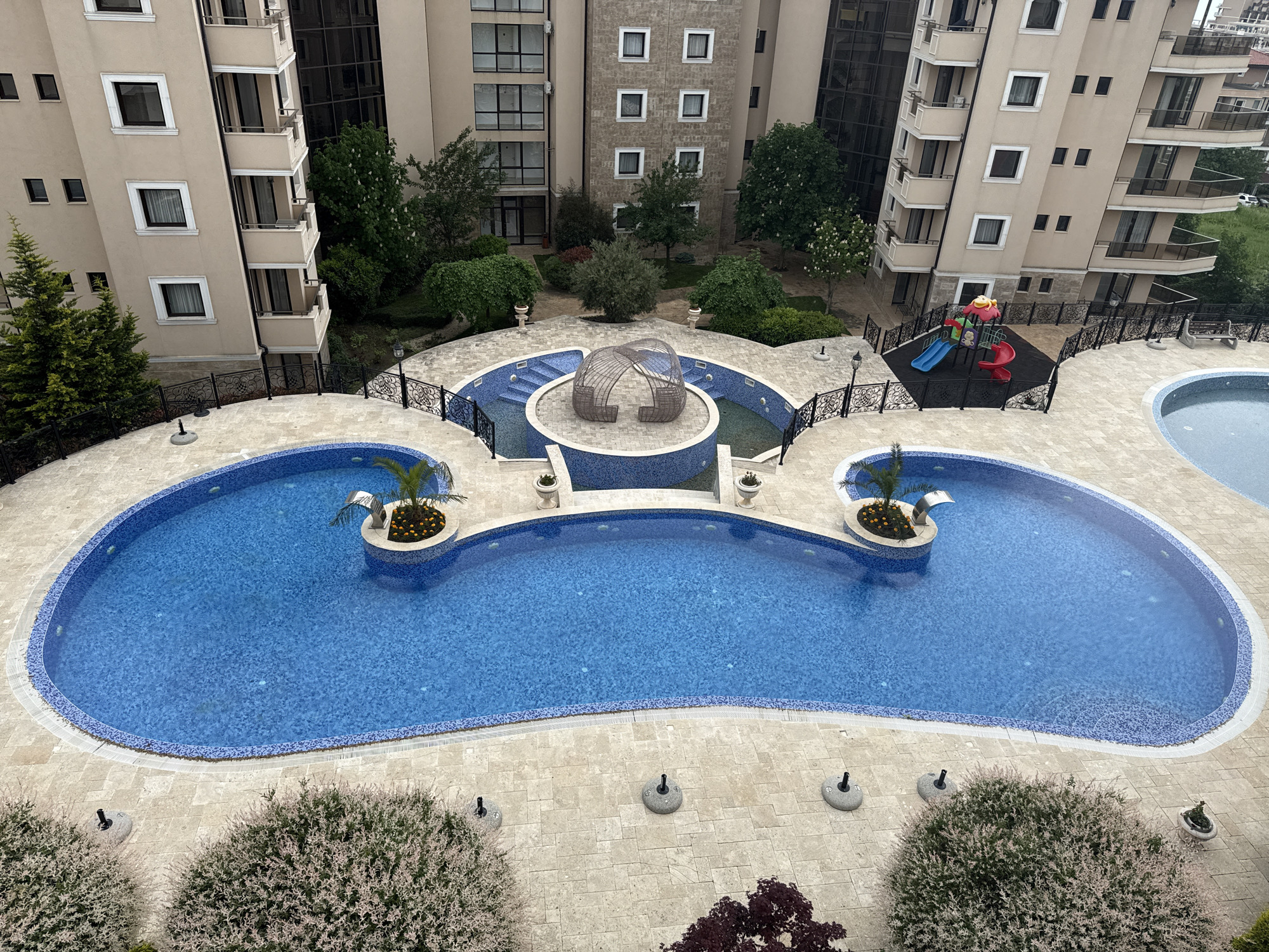 This photograph, likely taken from a high vantage point, showcases a resort-style pool complex in Sveti Vlas, Bulgaria. The complex features a large, kidney-shaped swimming pool with blue-tiled interior and bordering. A couple of small, elevated islands are centered within the pool, each landscaped with plants and a fountain. Adjacent to the main pool is a shallow wading pool or decorative water feature with a central wicker gazebo-like structure, surrounded by landscaped areas. To the upper right is a playground with colorful equipment, enclosed by a black fence. The area is surrounded by multi-story residential buildings with balconies. The foreground includes landscaped areas with small trees and shrubs. The weather appears overcast. No people are visible in the shot.