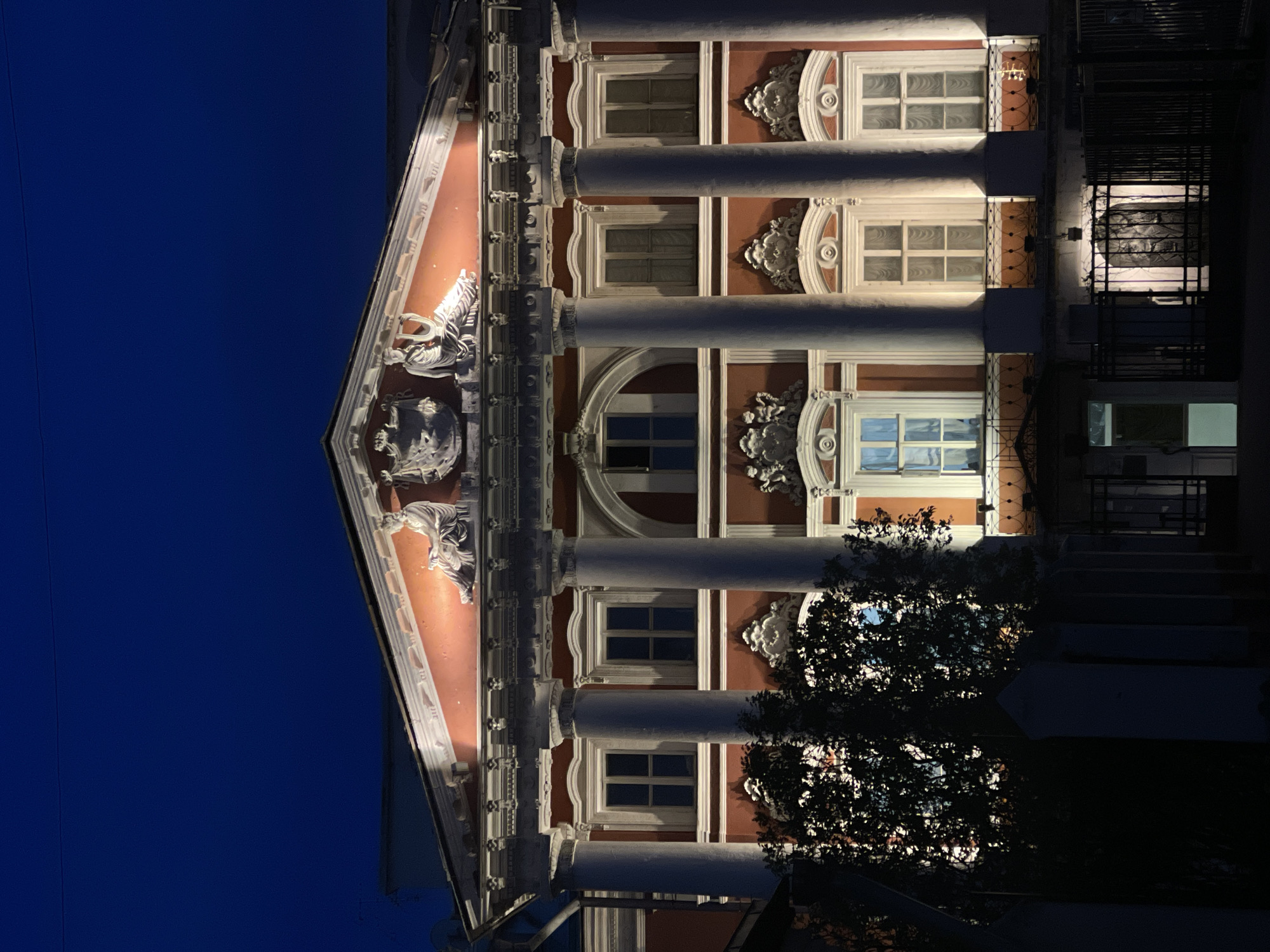 Here is a detailed description of the image:
**Scene:** The image depicts the facade of a building at night. The building is illuminated with lights, highlighting its architectural features.
**Architecture:** The building features classical architectural elements such as columns, windows, and decorative details. The facade is decorated with sculptures and moldings. The pediment is decorated with sculptural figures.
**Environment:** The sky is a dark blue color, suggesting that it is either late evening or night. The presence of trees indicates that the building is located in an area with vegetation.
**Location:** The building is identified as the Manezh Central Exhibition Hall in Moscow, Russia, based on the architectural style and the presence of recognizable elements.