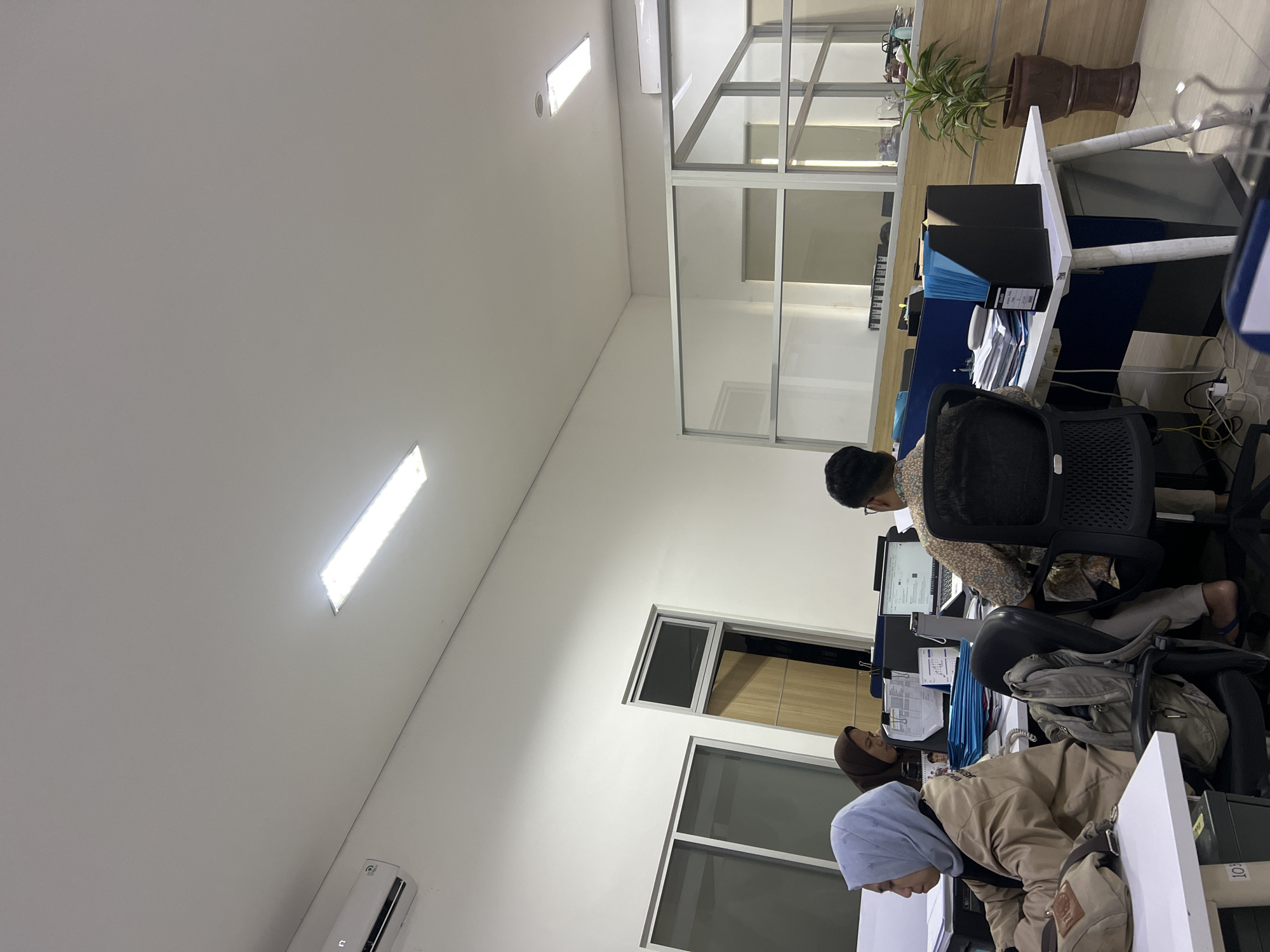 The image depicts an indoor office environment, likely in Yogyakarta, Indonesia, based on the context provided. The scene shows several individuals working at their desks. In the foreground on the left, a person wearing a light blue hijab and a beige jacket is seated at a white desk, looking at a computer. Next to them, another person in a brown hijab is also seated at a desk. Further to the right, a person wearing a patterned shirt is looking at a laptop. The office is equipped with modern amenities, including fluorescent ceiling lights and an air conditioning unit. The workspace is divided by glass partitions, suggesting a semi-open office plan. Desks are cluttered with office supplies such as papers, folders, and electronics. A potted plant is visible in a brown pot on a shelf in the background. The overall impression is one of a functional, contemporary office space.