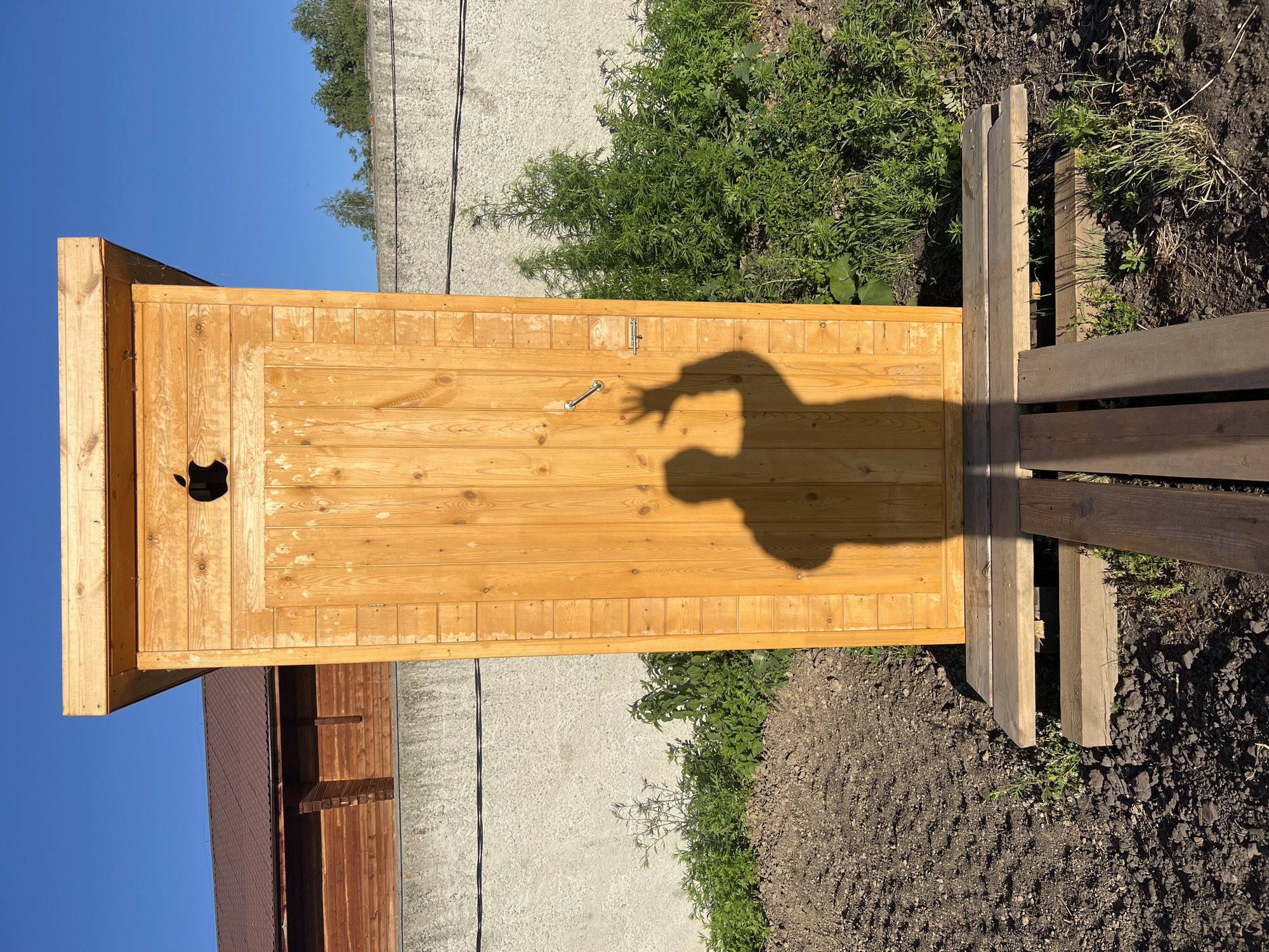 A wooden outhouse stands in the foreground with its door ajar, casting a shadow of a person with a raised hand. The outhouse is positioned on a wooden platform, with planks leading to it from the ground. Around the outhouse, dark soil is visible, with patches of green grass and small plants growing. In the background, a grey concrete wall runs horizontally across the image, with a wooden building and trees behind it. The sky is a clear, bright blue. The scene appears to be in a rural setting, likely a backyard or garden, with elements suggesting a home in Novopichugovo, Russia.