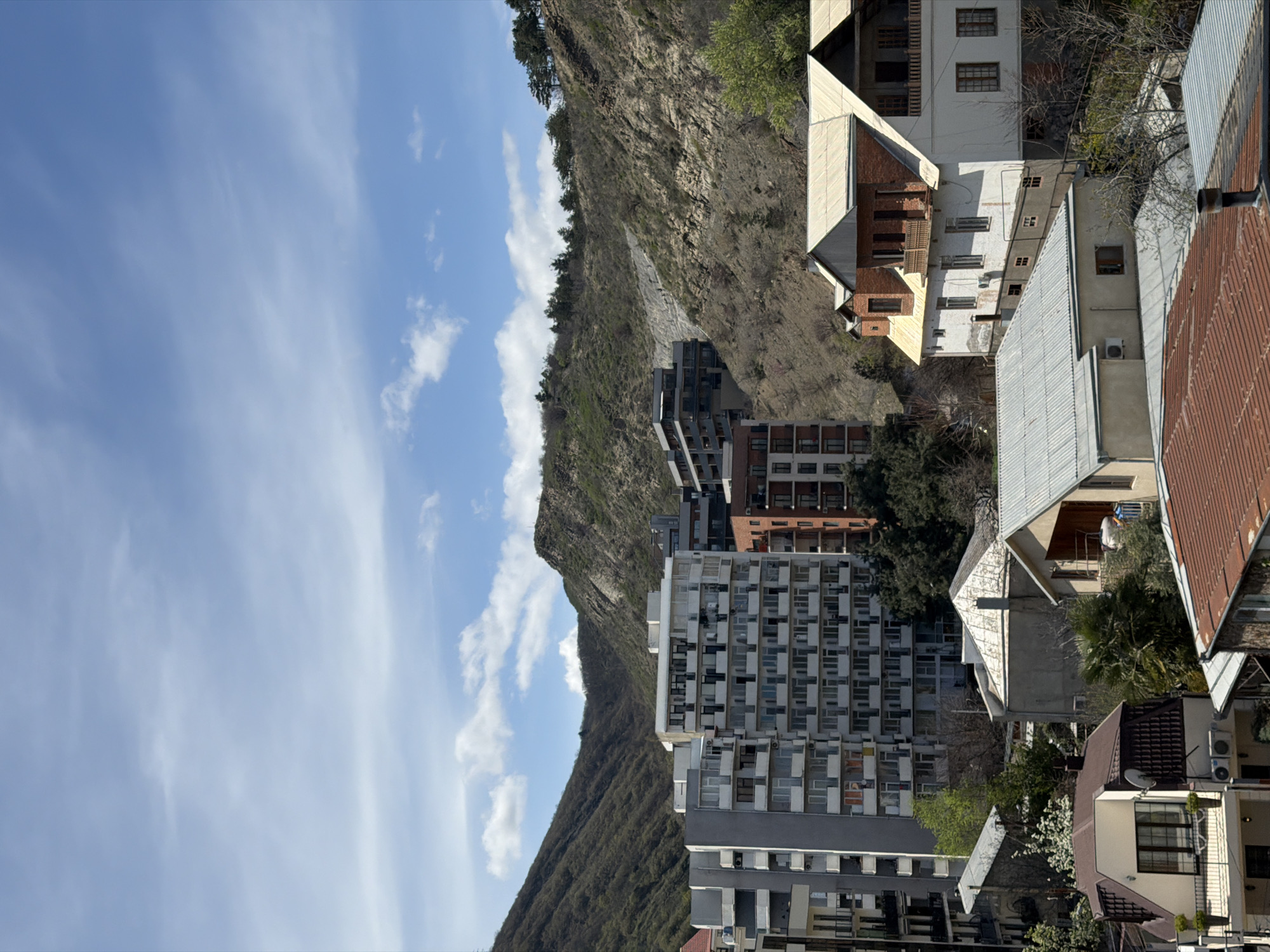 A low-angle shot showcases a cityscape in Tbilisi, Georgia, featuring a variety of buildings and the presence of a mountain landscape. In the background, a mountain is visible, partially covered in vegetation and rock formations. The sky, predominantly blue, contains scattered white clouds.
The architecture in the scene includes a mix of high-rise apartment buildings and individual houses with pitched roofs. The apartments vary in color and style. The houses have roofs made of different materials, including metal and tile. Some residences are equipped with air conditioning units.
Visible are trees, some in early blossom. The scene is bathed in daylight. No people are visible in the image.