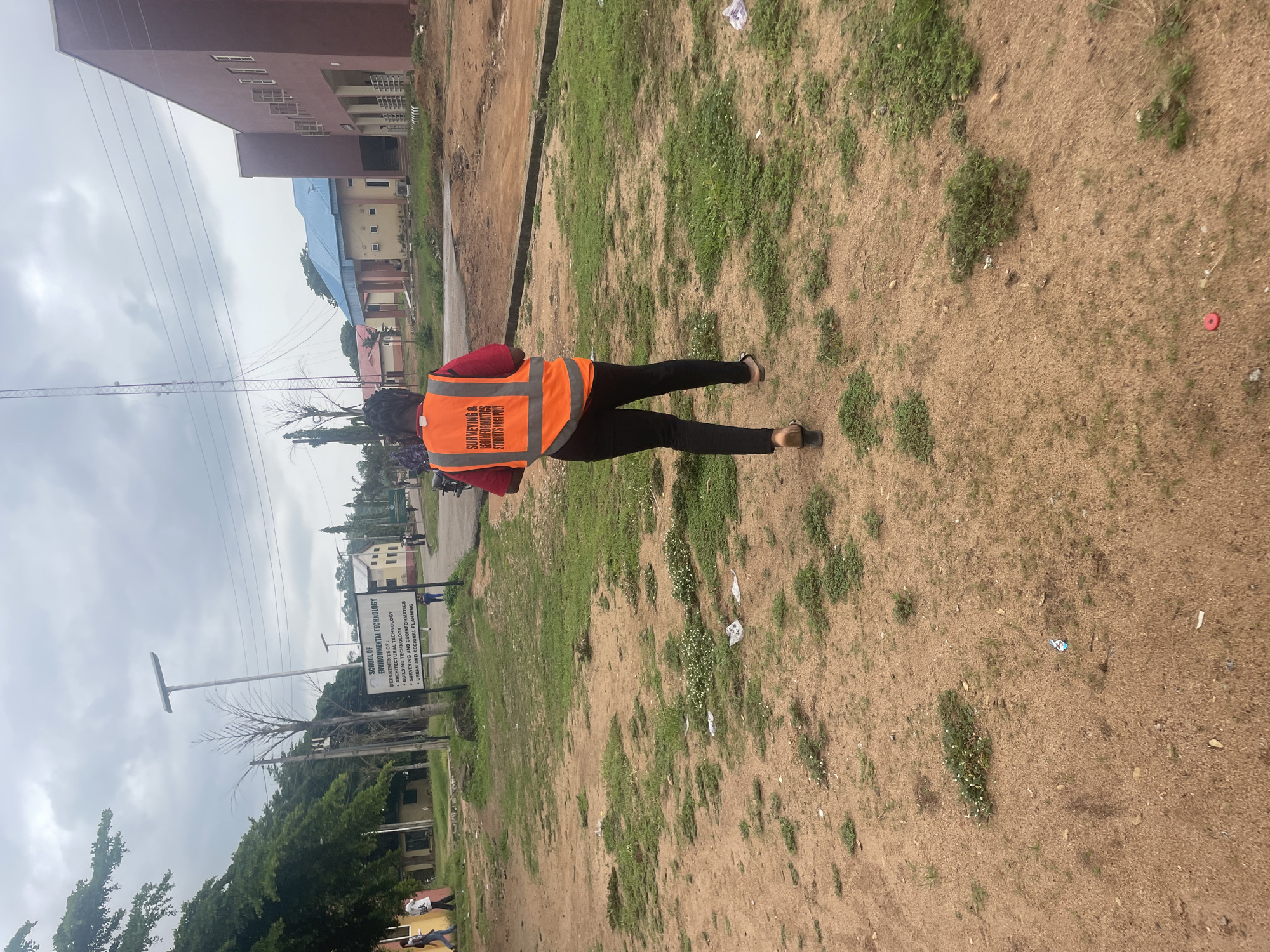 The image shows a person wearing an orange safety vest over a red shirt and black pants, walking away from the camera on a dirt and grass pathway. The vest has text that reads "SURVEYING & GEOMATICS STUDENTS KOGI POLY". The background features buildings with red and blue roofs, trees, a cell tower, and a sign for "SCHOOL OF ENVIRONMENTAL TECHNOLOGY". The environment appears to be outdoors, possibly on a campus in Lokoja, Nigeria, indicated by the "KOGI POLY" text. The sky is overcast. The ground is uneven, with patches of dirt and grass, and some scattered litter. There are a few other people visible in the distance, near the buildings. The overall impression is one of an academic or administrative setting.