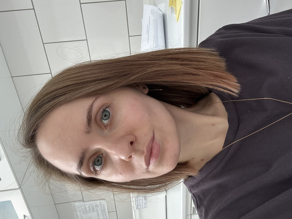 A fair-skinned woman with light brown, shoulder-length hair is shown in a close-up selfie. She has striking blue eyes and defined eyebrows. She wears a plain, dark-colored shirt and a delicate gold necklace. The background consists of white tiles, suggesting a bathroom or kitchen setting, with some papers partially visible in the top corner. The image is slightly rotated.