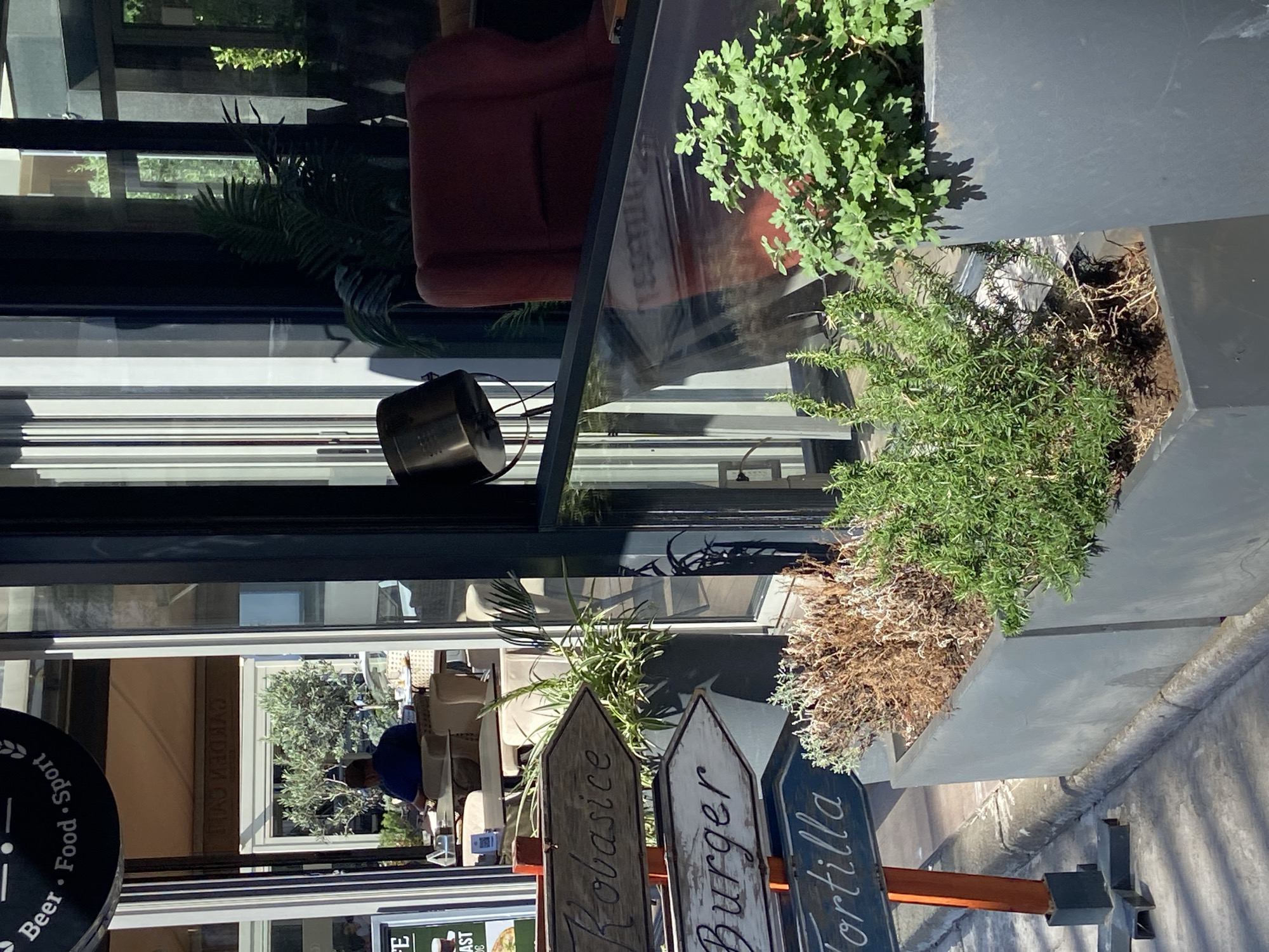 The image shows an outdoor scene in Budva, Montenegro, featuring a restaurant or cafe. The exterior includes potted plants and directional signs advertising "Klobasice," "Burger," and "Tortilla." Through the glass windows, a person is seen sitting at a table inside the establishment. The facade reflects the street scene, including a red chair and further vegetation. A sign in the lower left advertises "Beer - Food - Sport."