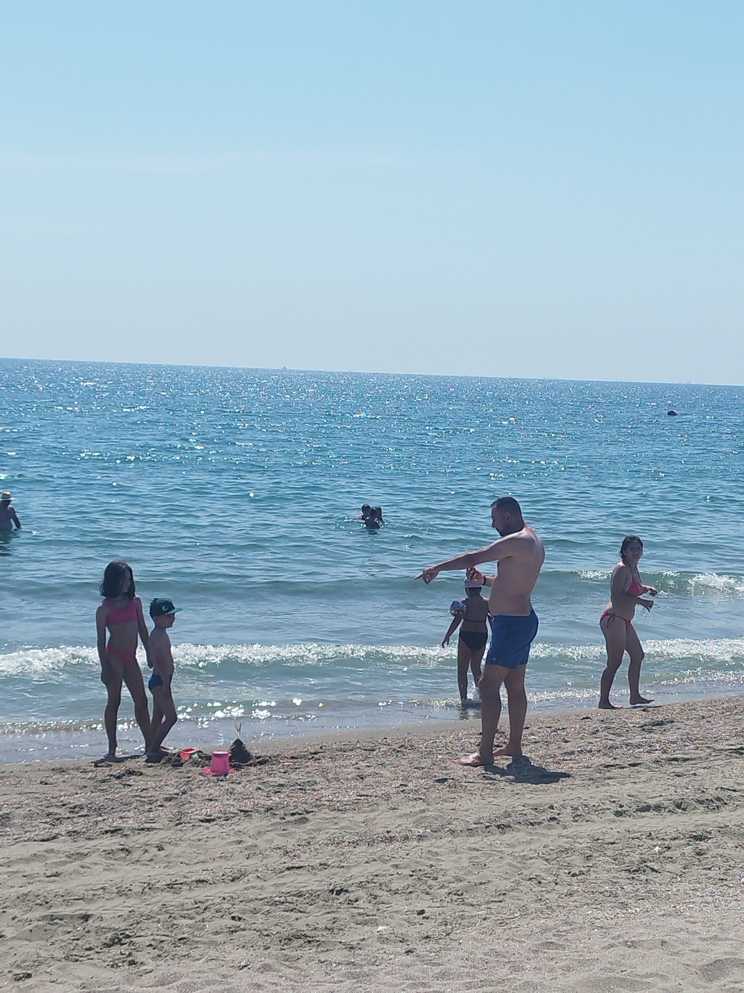 This image captures a sunny day at the beach in Constanța, Romania. Several people are enjoying the water and the shore. In the foreground, a young girl in a pink bikini and a boy in blue swim trunks stand near the water's edge, with beach toys scattered on the sand behind them. Further out in the sea, a man in blue swim trunks gestures towards the water, while a woman in a red bikini stands to his right. Other swimmers are visible in the middle ground, with some appearing to be children. The ocean is calm with gentle waves, reflecting the bright sunlight. The sky is clear and blue, indicating fair weather. The sandy beach is covered in small shells and pebbles. The overall scene suggests a leisurely summer day spent by the sea.