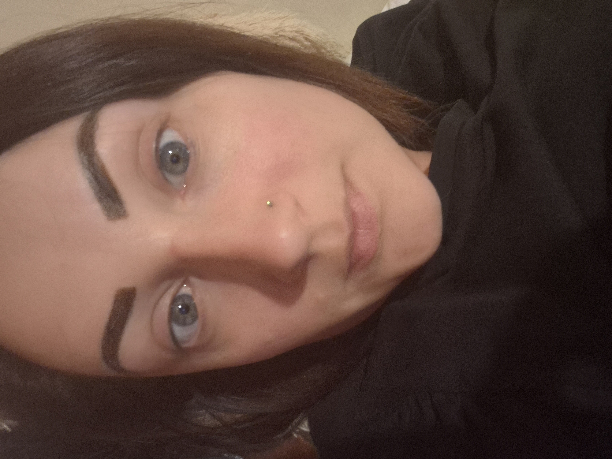 The image is a close-up selfie of a woman with dark brown hair and light blue eyes. She is wearing a black top. She has on dark, defined eyebrows and a small stud piercing in her right nostril. Her skin is fair with some redness on her cheeks. The lighting appears to be indoor. The image does not provide any information about the location within the city of Sandwell, United Kingdom.