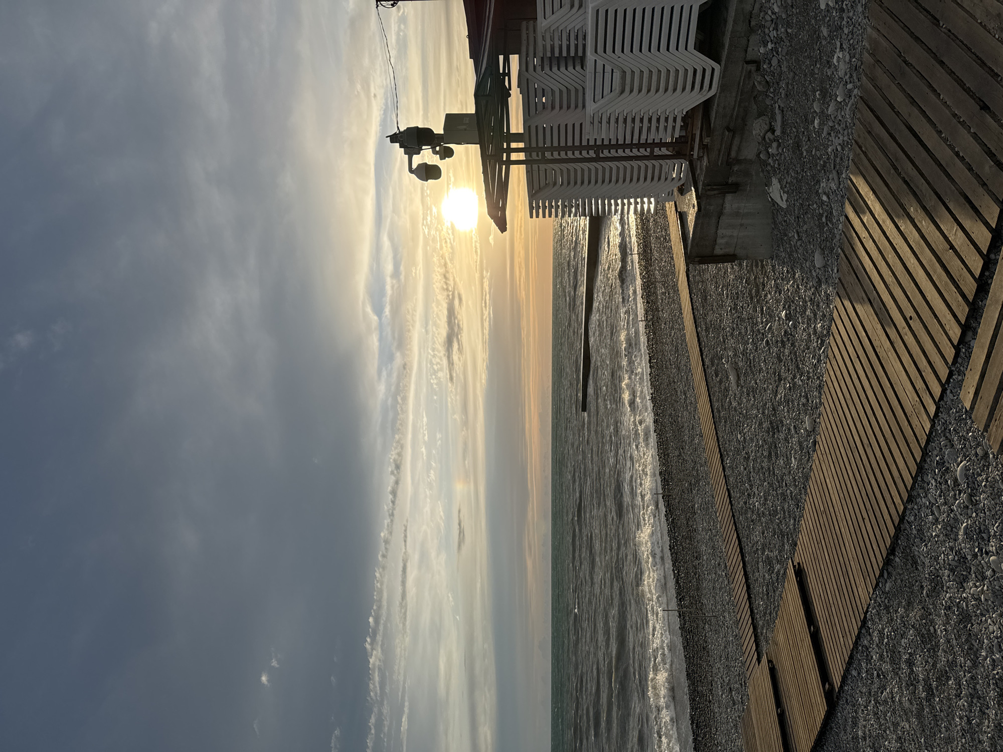 The image shows a sunset on the beach in Sochi, Russia. The sun is setting over the Black Sea, casting a golden light across the water and the sky. On the right side of the image, there is a wooden boardwalk, a section of pebble beach and a stack of white beach chairs. A weather monitoring station can be seen just to the left of the stack of chairs. The sky is filled with clouds. The sea is calm, with gentle waves lapping at the shore.